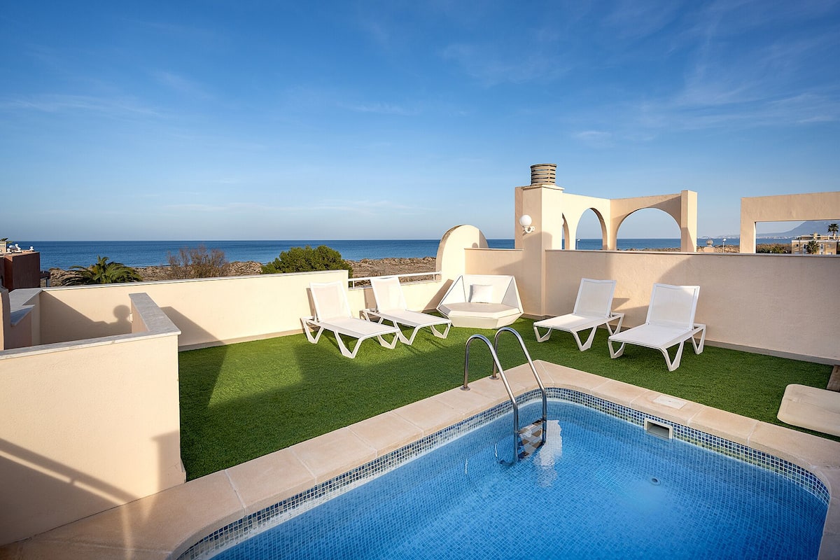 Une piscine sur le toit offrant une vue dégagée sur l’océan, entourée de chaises longues blanches et d’un petit espace aménagé au bord de la piscine.