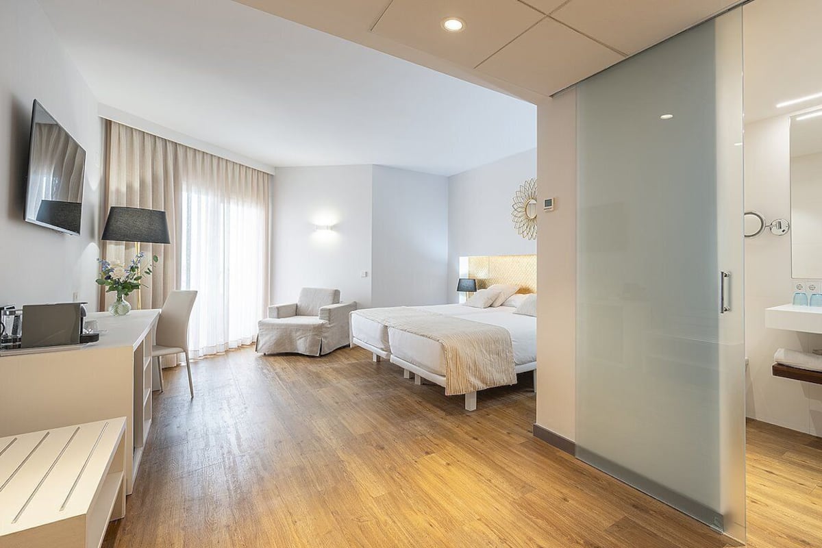 Une chambre d’hôtel moderne avec un plancher en bois, un lit, un bureau avec une chaise, une télévision et une salle de bain équipée d’un miroir et d’un lavabo.