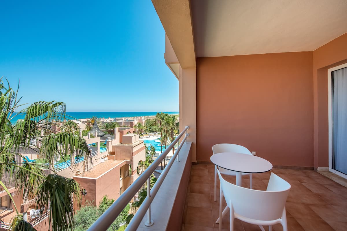 Un balcon donnant sur une piscine et des palmiers, une table et une chaise blanches, et un mur de couleur pêche.