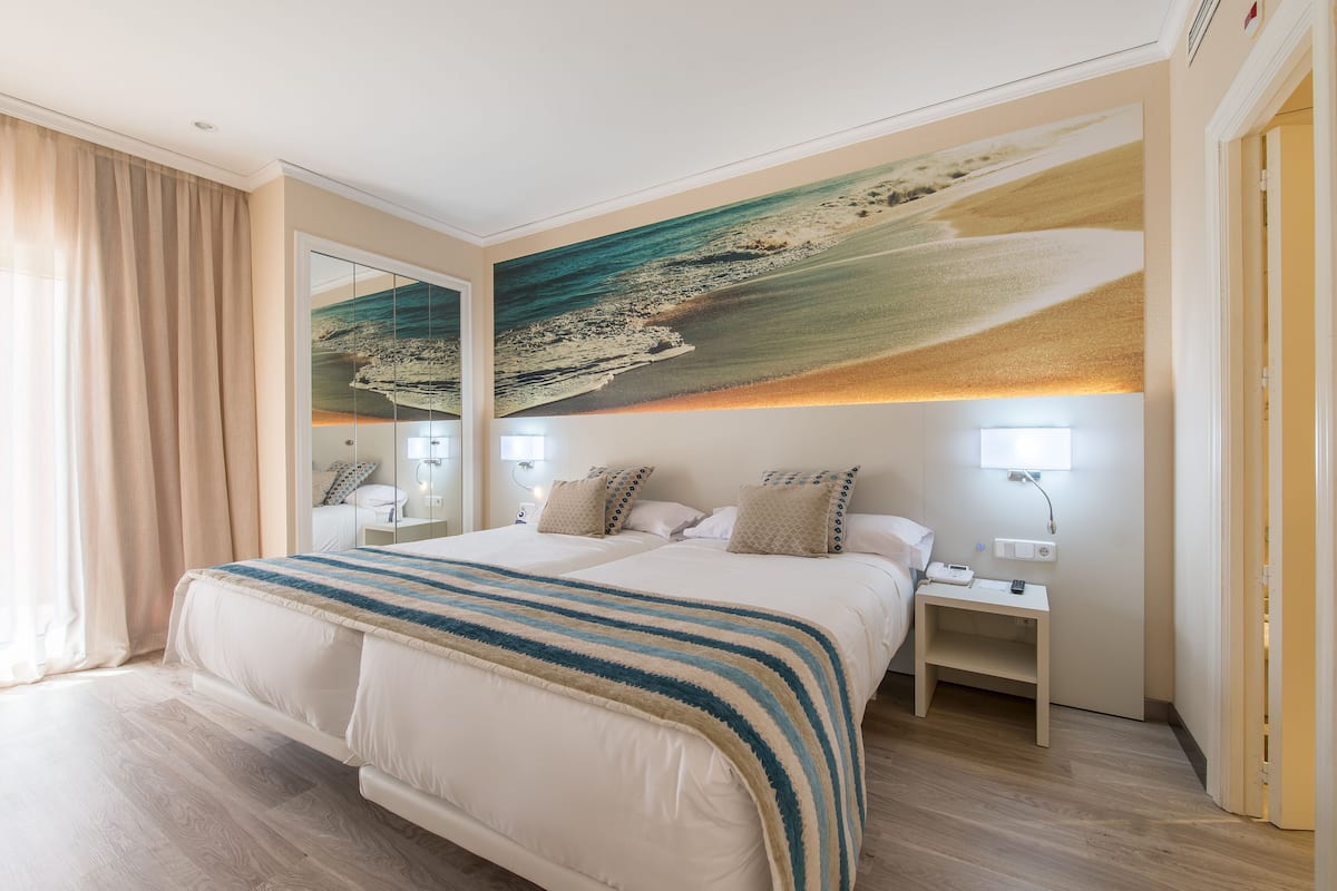 Une chambre d’hôtel avec un grand lit, une table de chevet, une lampe fixée au mur et un grand tableau représentant une scène de plage.
