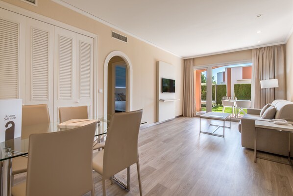 Une chambre d’hôtel moderne avec une table à manger en verre, des chaises beiges, un canapé et un téléviseur fixé au mur.