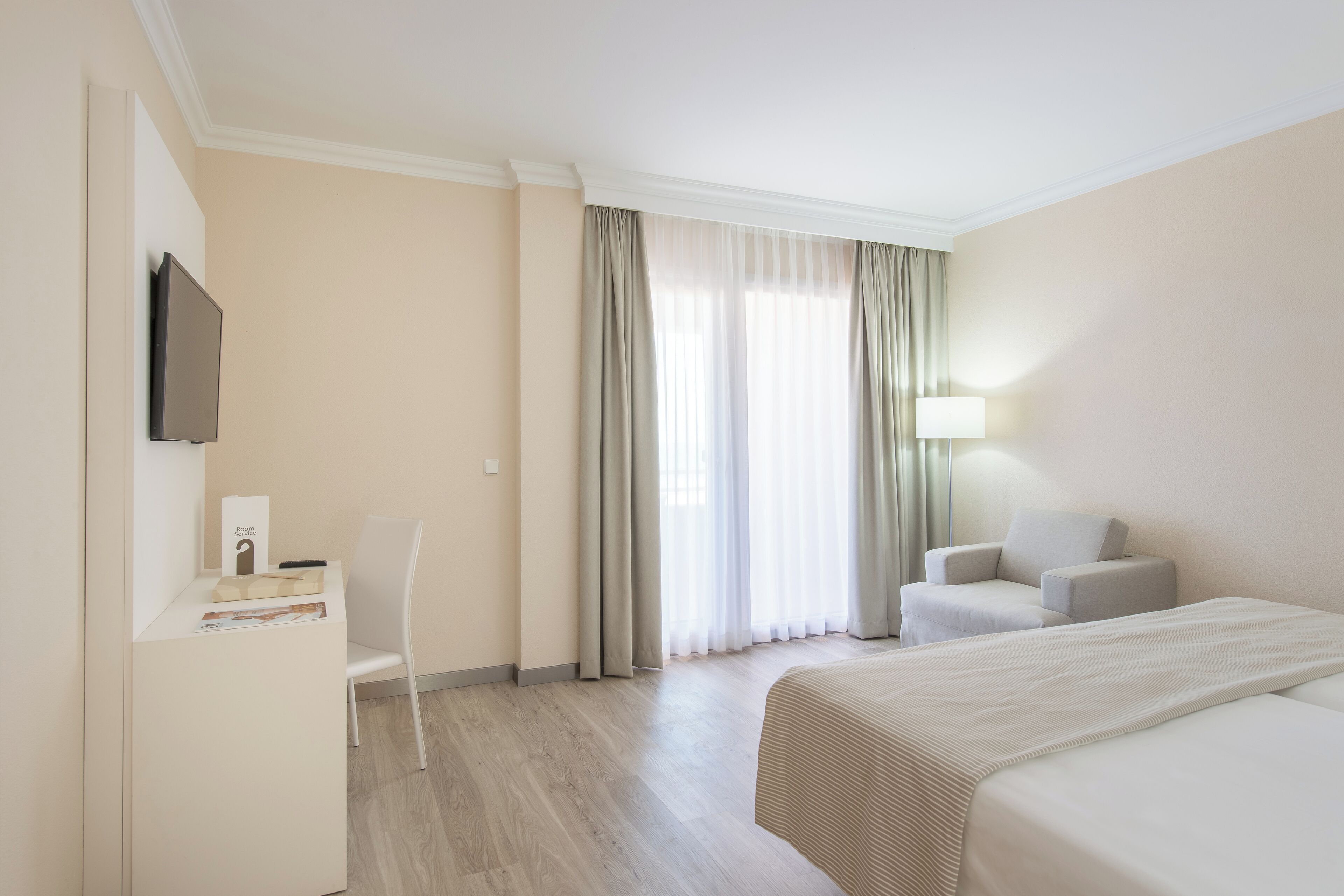 A hotel room with a bed, a desk with a chair, a TV, and a window with curtains.