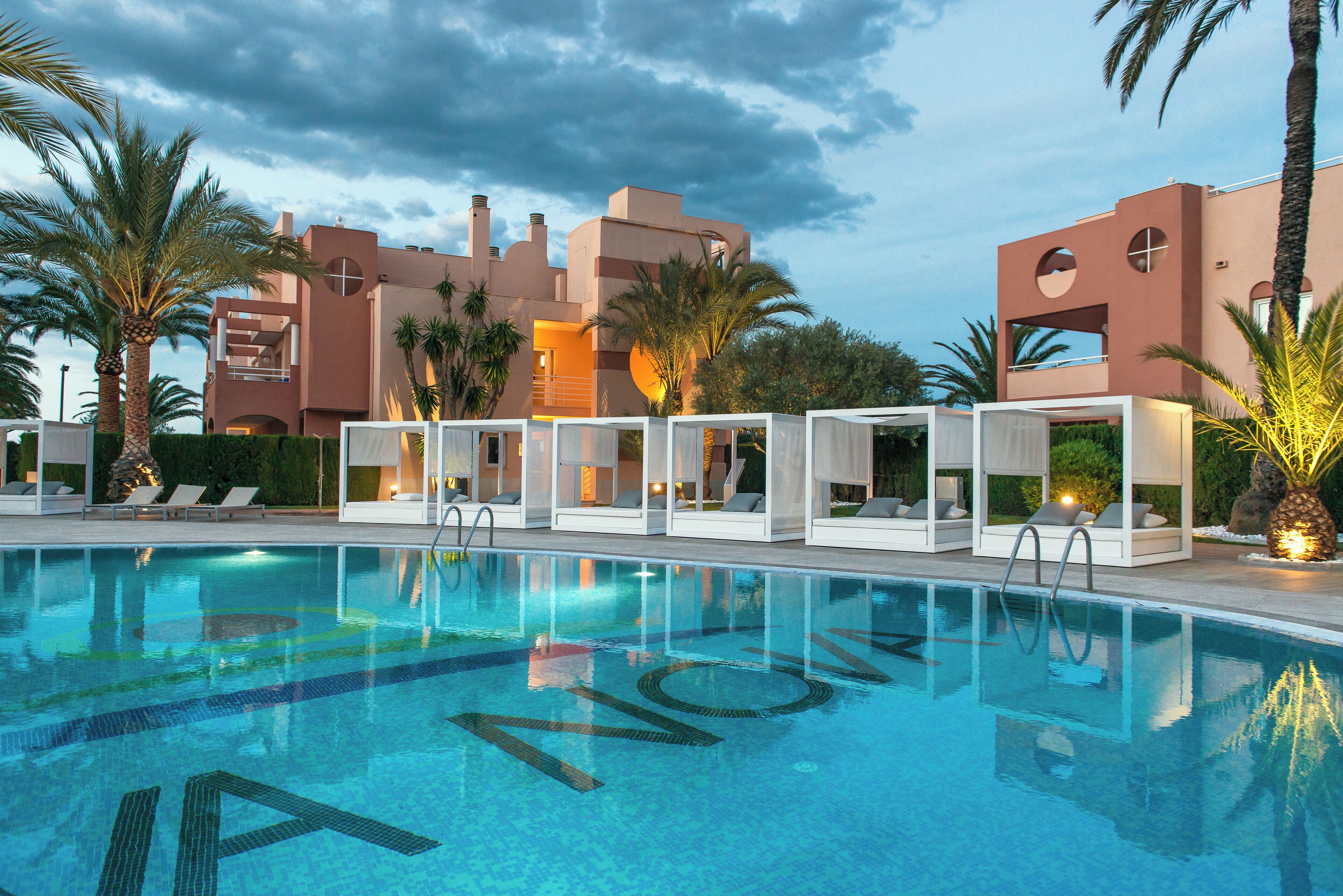 A swimming pool with a clear view of the poolside lounge area, palm trees, and modern buildings in the background.
