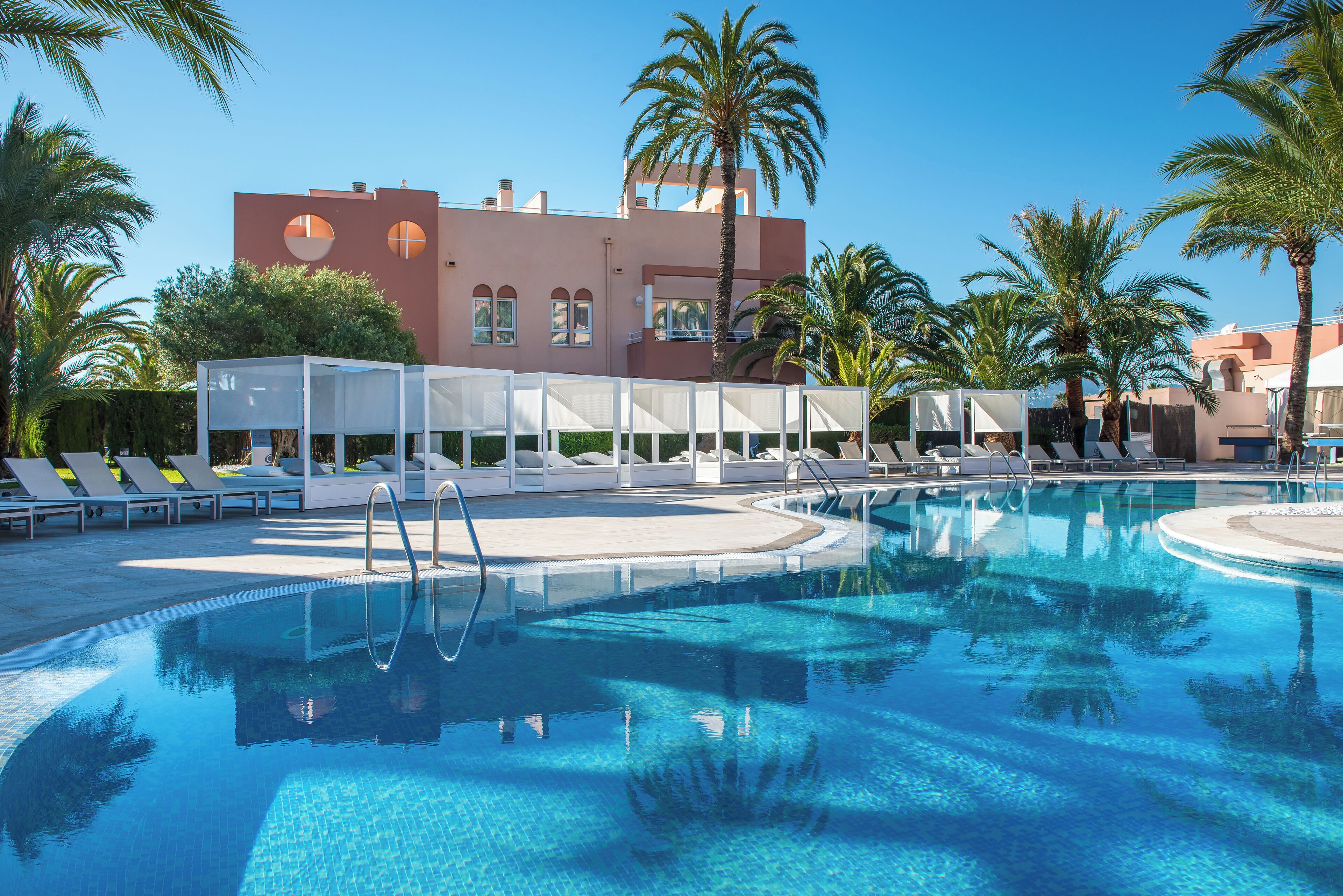 Una piscina con sillas de descanso y sombrillas, rodeada de palmeras y un edificio con grandes ventanales.