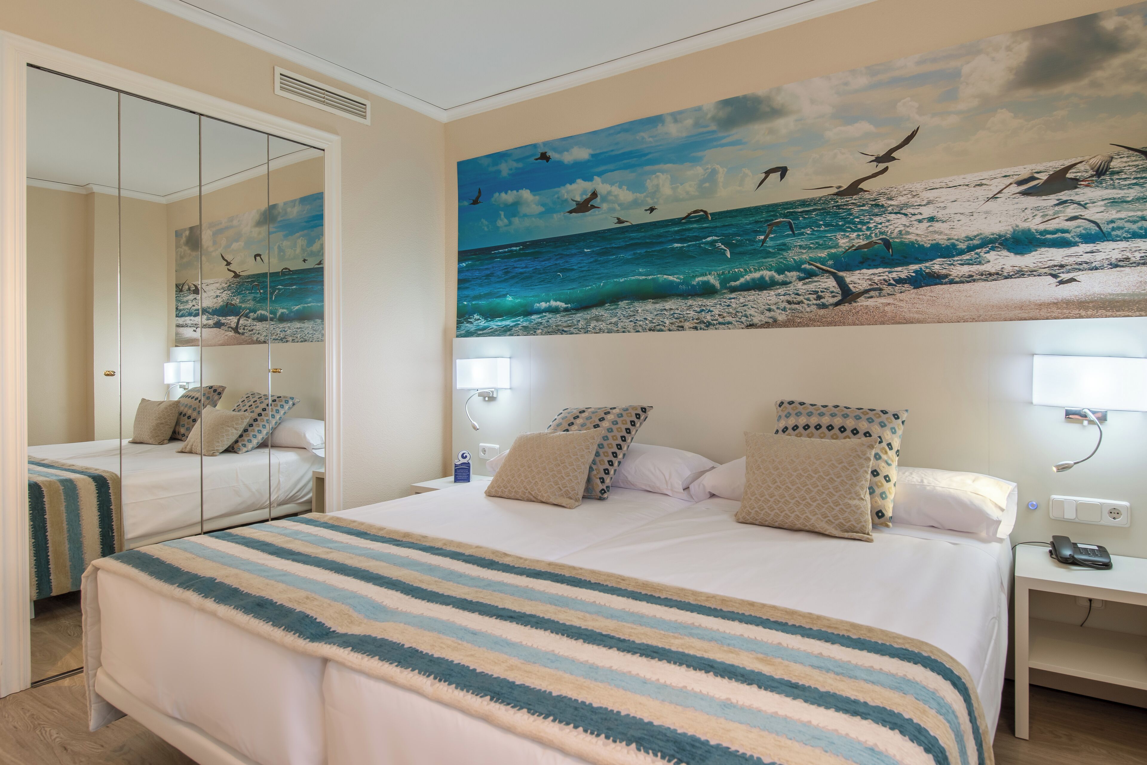 A hotel room with a large bed, a bedside table, a mirror, and a wall mural of a beach scene.