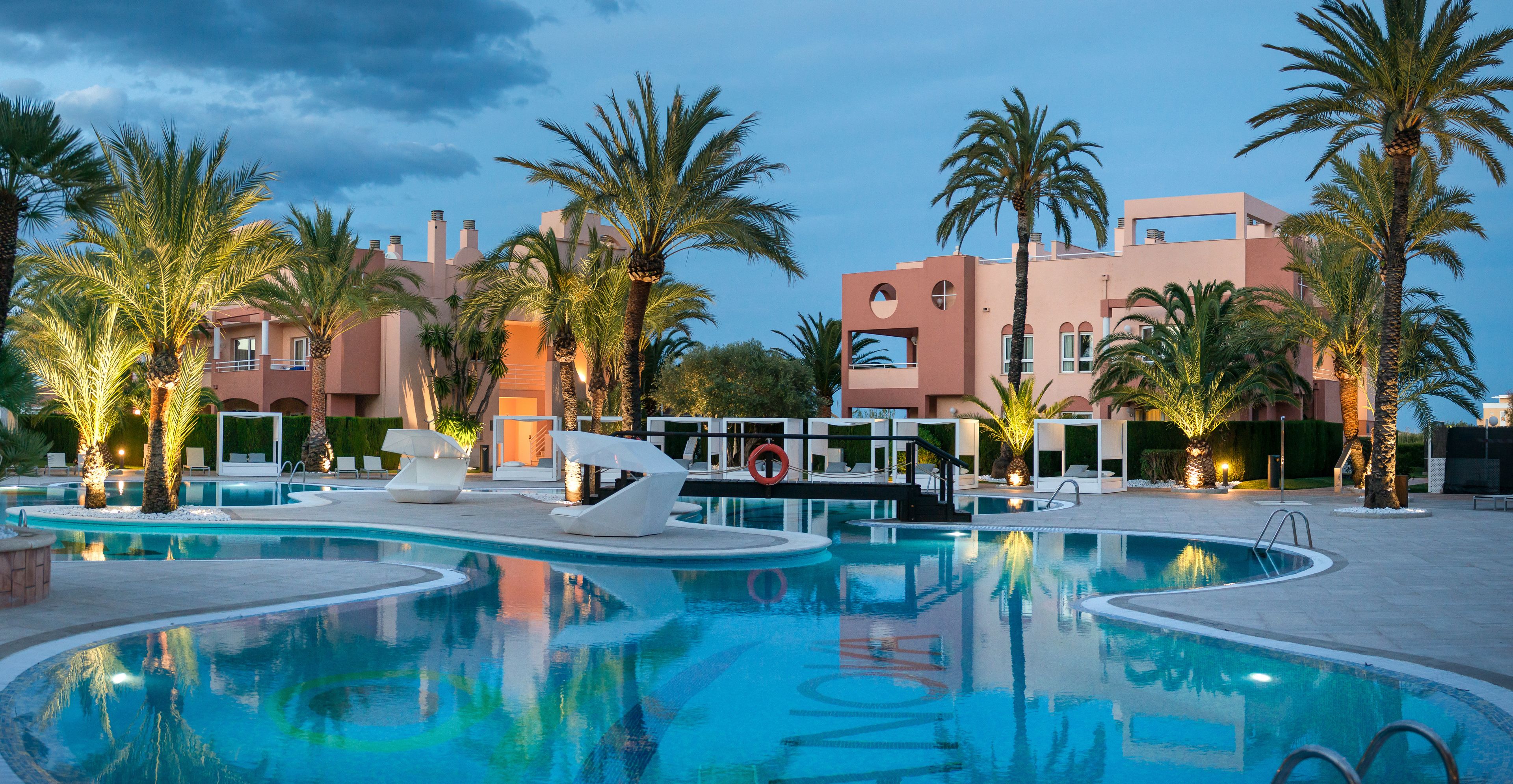 Área de piscina en un resort con palmeras, sillas de descanso y un edificio moderno con balcones.