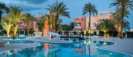 Área de piscina en un resort con palmeras, sillas de descanso y un edificio moderno con balcones.