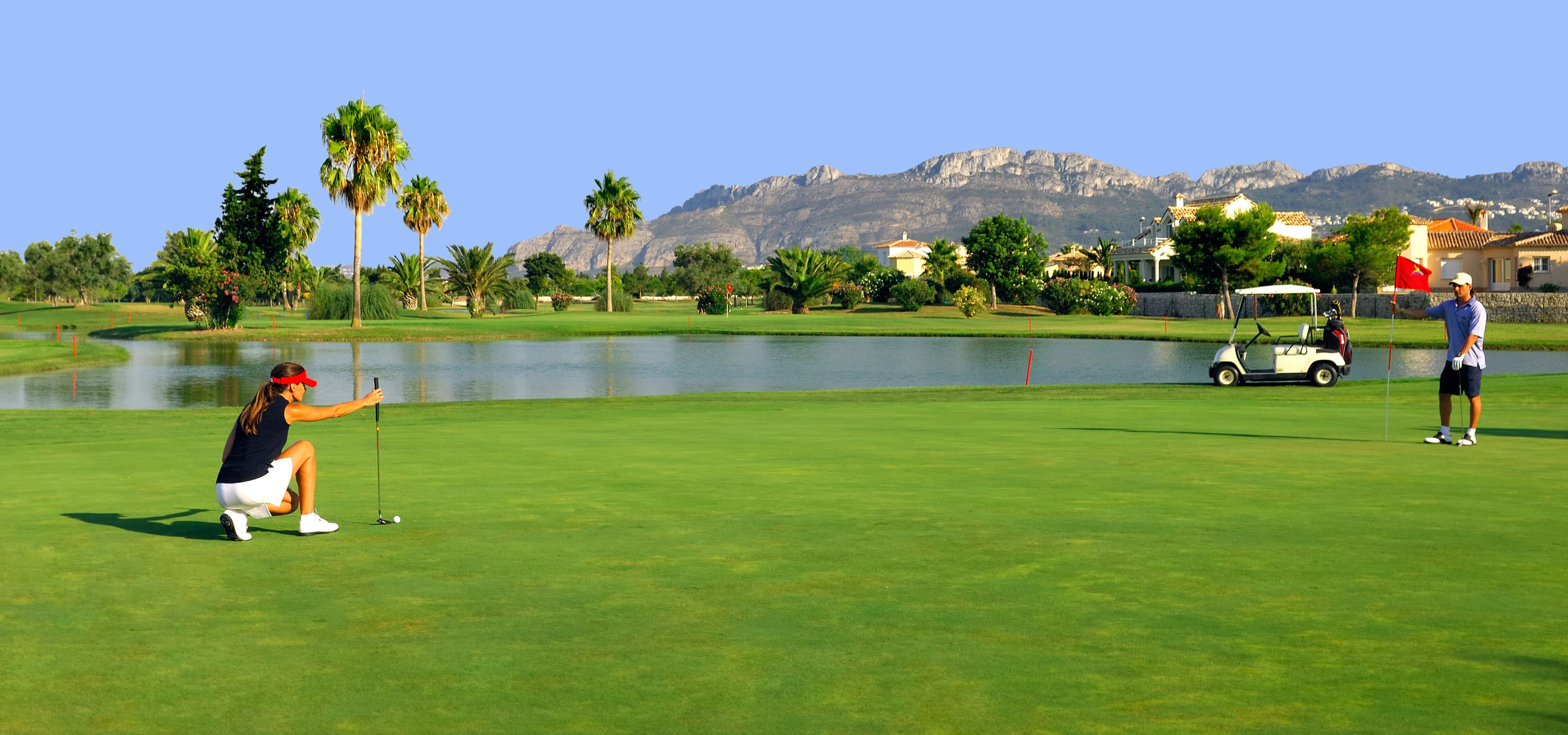 Un campo de golf con un lago, palmeras y montañas al fondo.