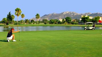 Un campo de golf con un lago, palmeras y montañas al fondo.
