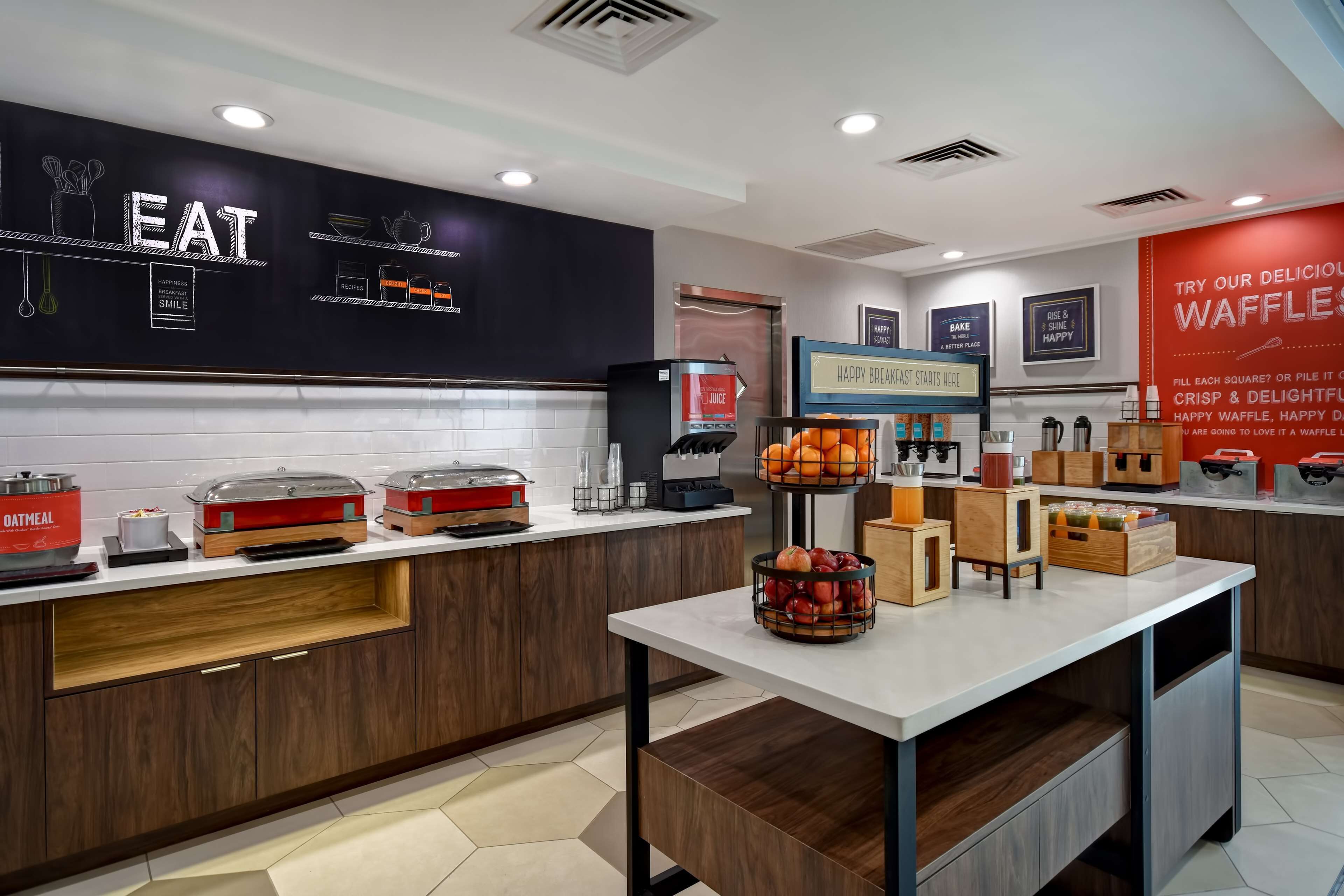 A modern kitchen area with a black wall displaying the word "EAT" and various kitchen utensils. There are food dispensers, a coffee machine, and a sign that says "HAPPY BREAKFAST STARTS HERE".