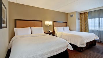 A hotel room with two beds, a nightstand with a lamp, a window with curtains, and a wall-mounted air conditioner.
