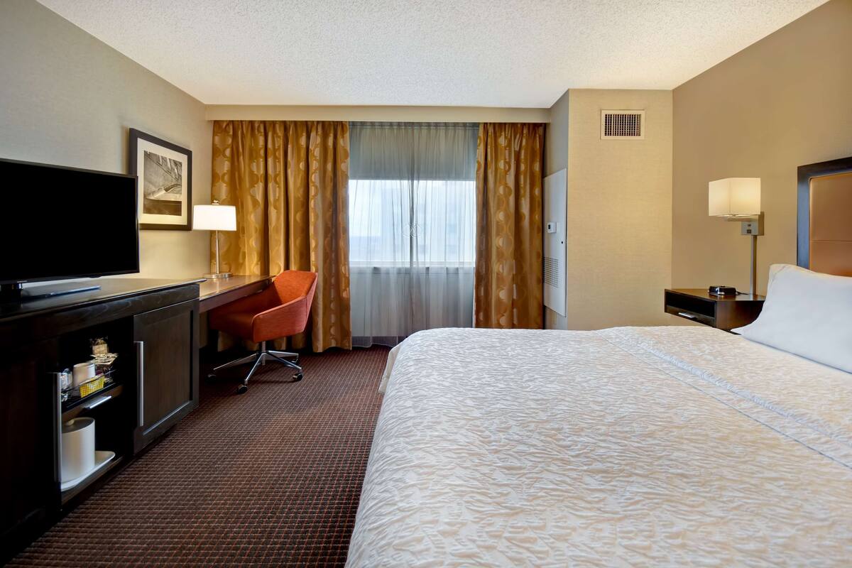 Ein Hotelzimmer mit einem großen Bett, einem Schreibtisch mit Flachbildfernseher, einem Sessel, einem Nachttisch mit Lampe und einem fenster mit Vorhängen.