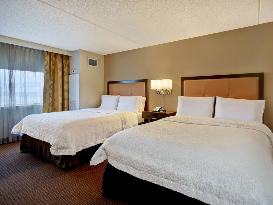 A hotel room with two beds, a nightstand with a lamp, a window with curtains, and a wall-mounted artwork.