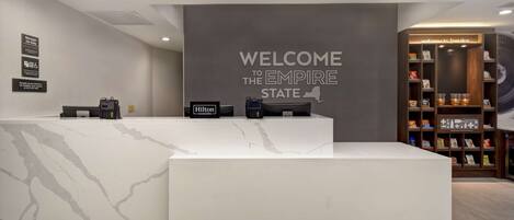 Zona de recepción de un hotel con mostrador de mármol, un cartel que dice 'Bienvenido al Empire State', un letrero que indica 'Gerente de Guardia', un cartel de 'Hilton Honors', y una estantería con aperitivos y una máquina de café.