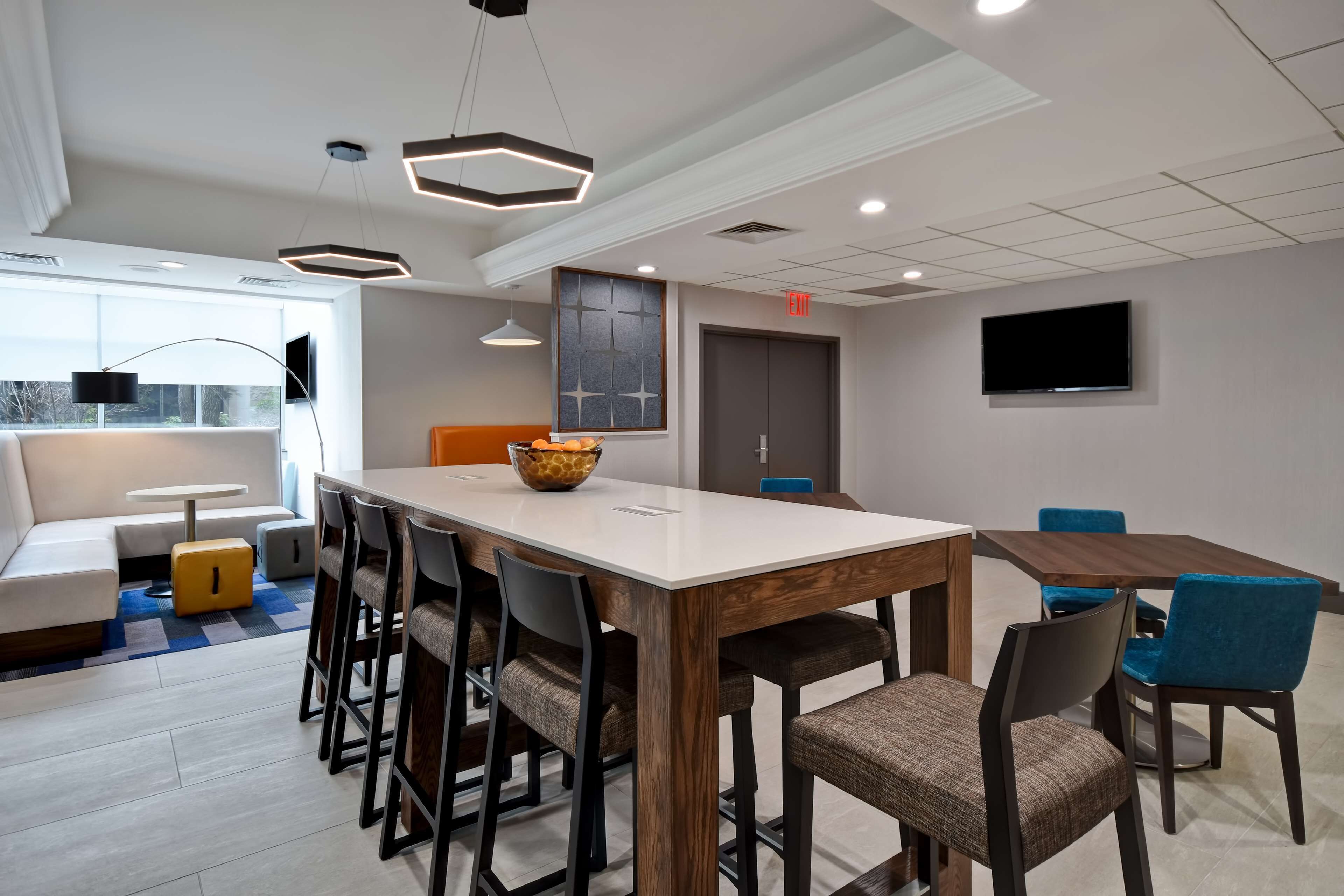 A modern hotel room with a large dining table, comfortable seating, a flat-screen TV, and a large window providing a view of the outdoors.