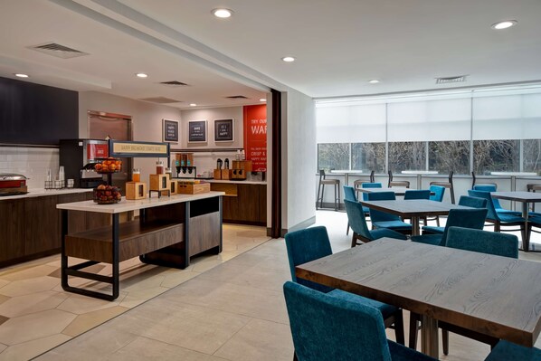 A modern breakfast area with a counter, seating, and a view of trees through large windows. - Hampton Inn NY-JFK (Jamaica)