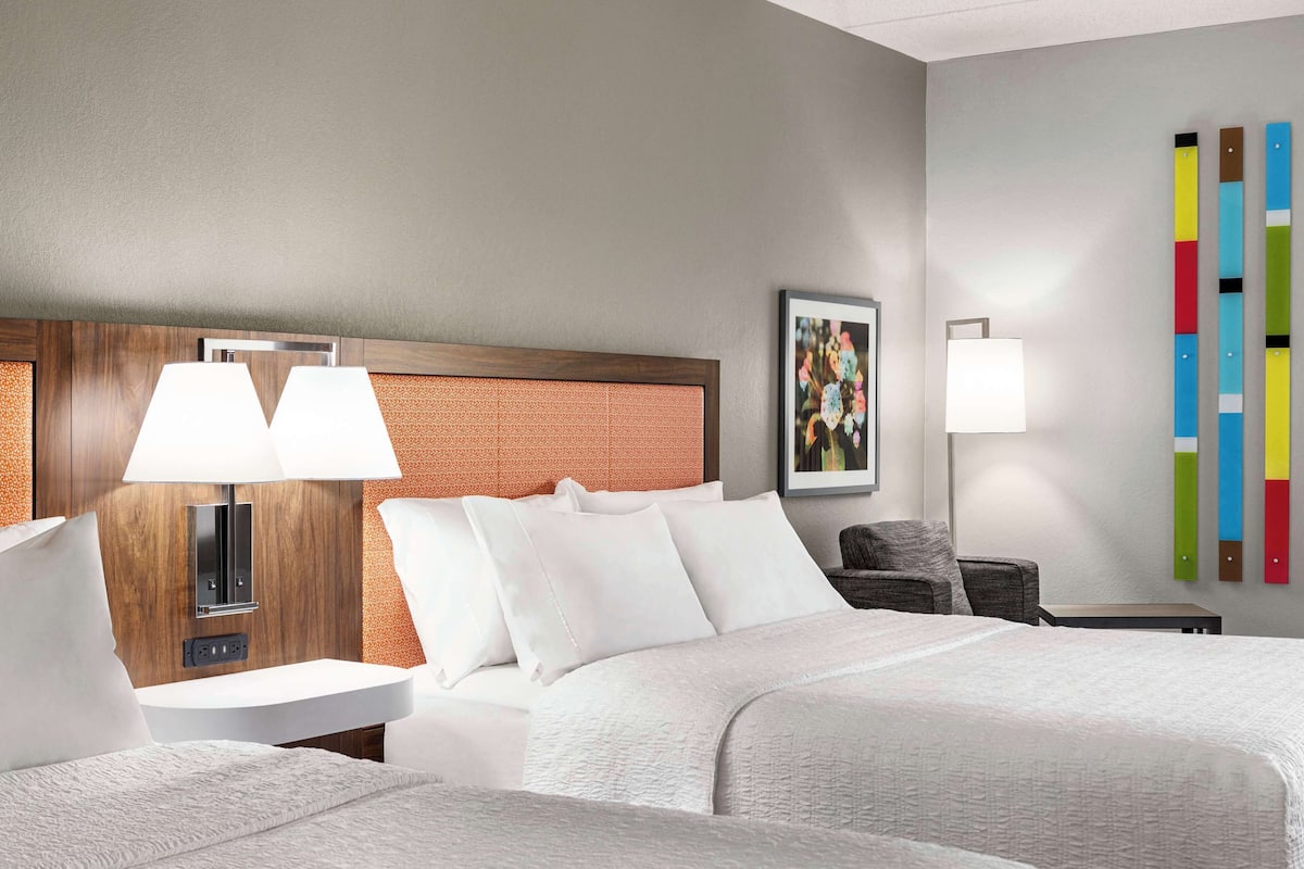 A hotel room with a large bed, a bedside table with a lamp, a wall-mounted TV, and a colorful abstract art piece.