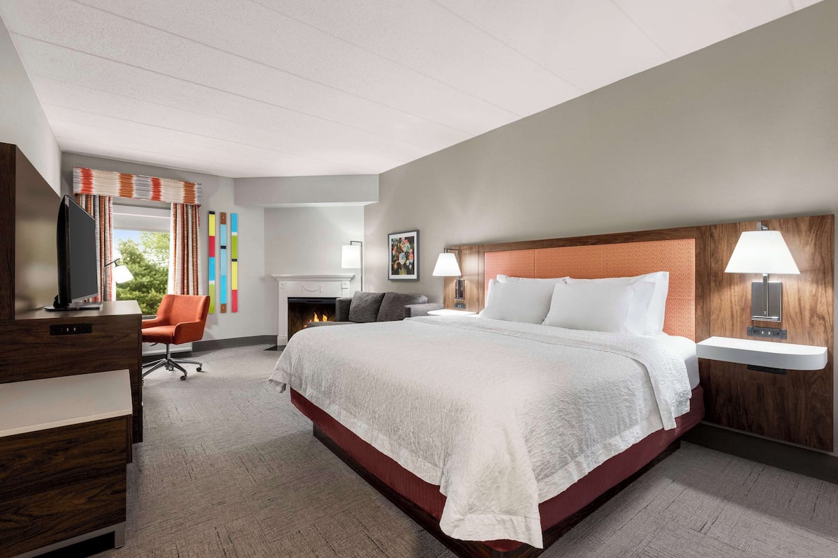 A hotel room with a large bed, a desk with a computer, a chair, a fireplace, and a window with curtains.
