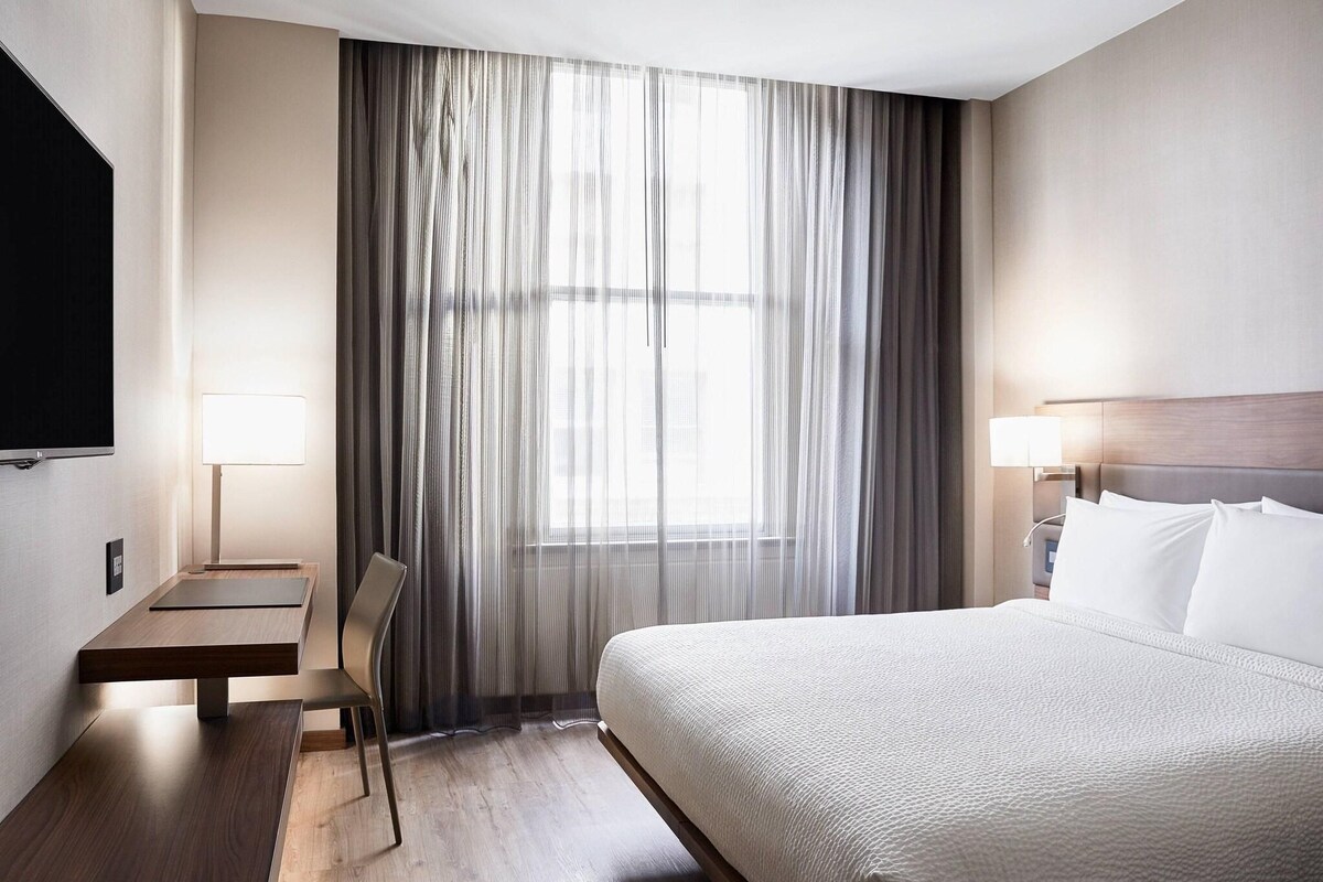 Une chambre d’hôtel avec un lit, un bureau, une chaise, une télévision et une fenêtre avec des rideaux.