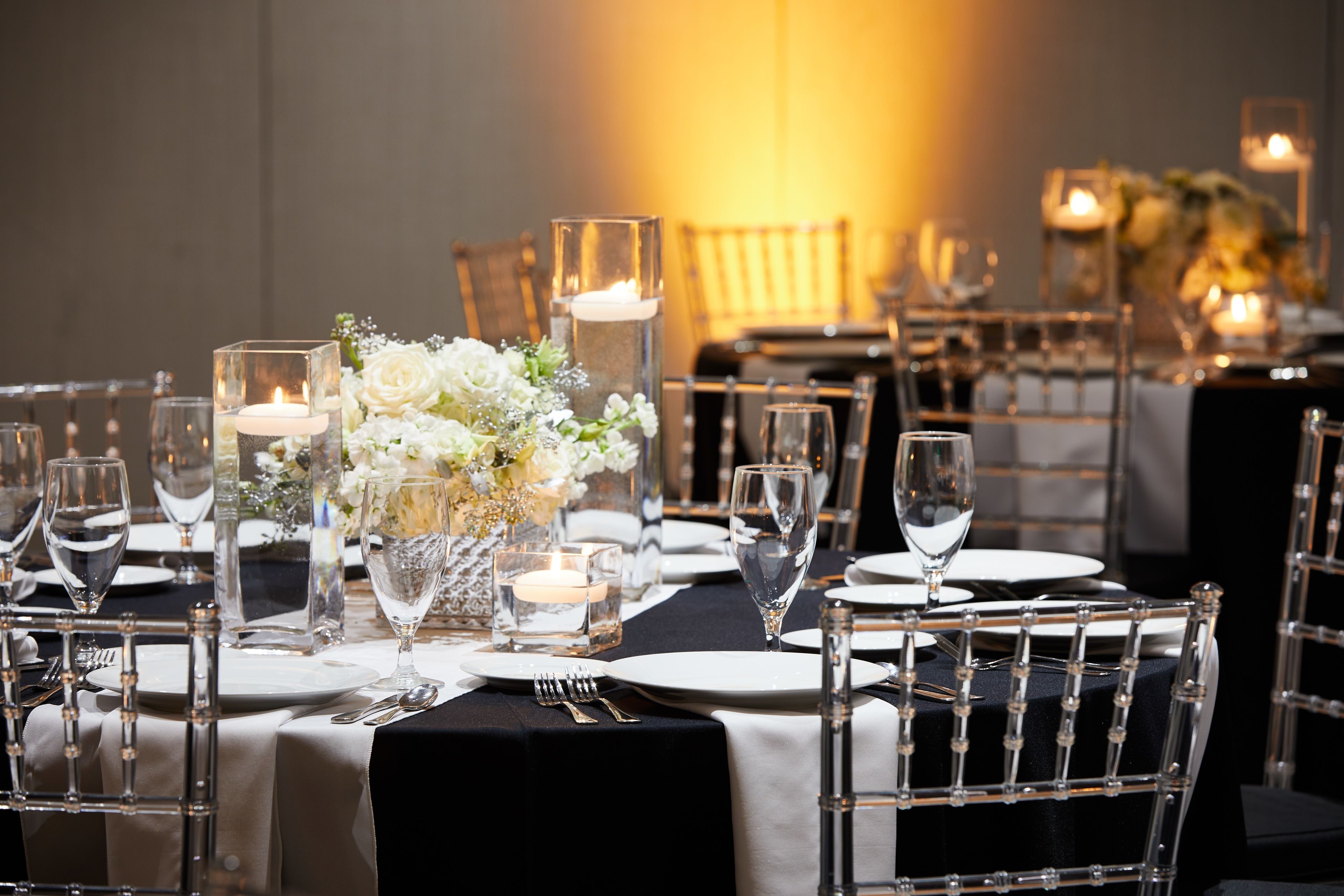 A dining table set with clear glassware, white plates, and a centerpiece of white flowers and candles.