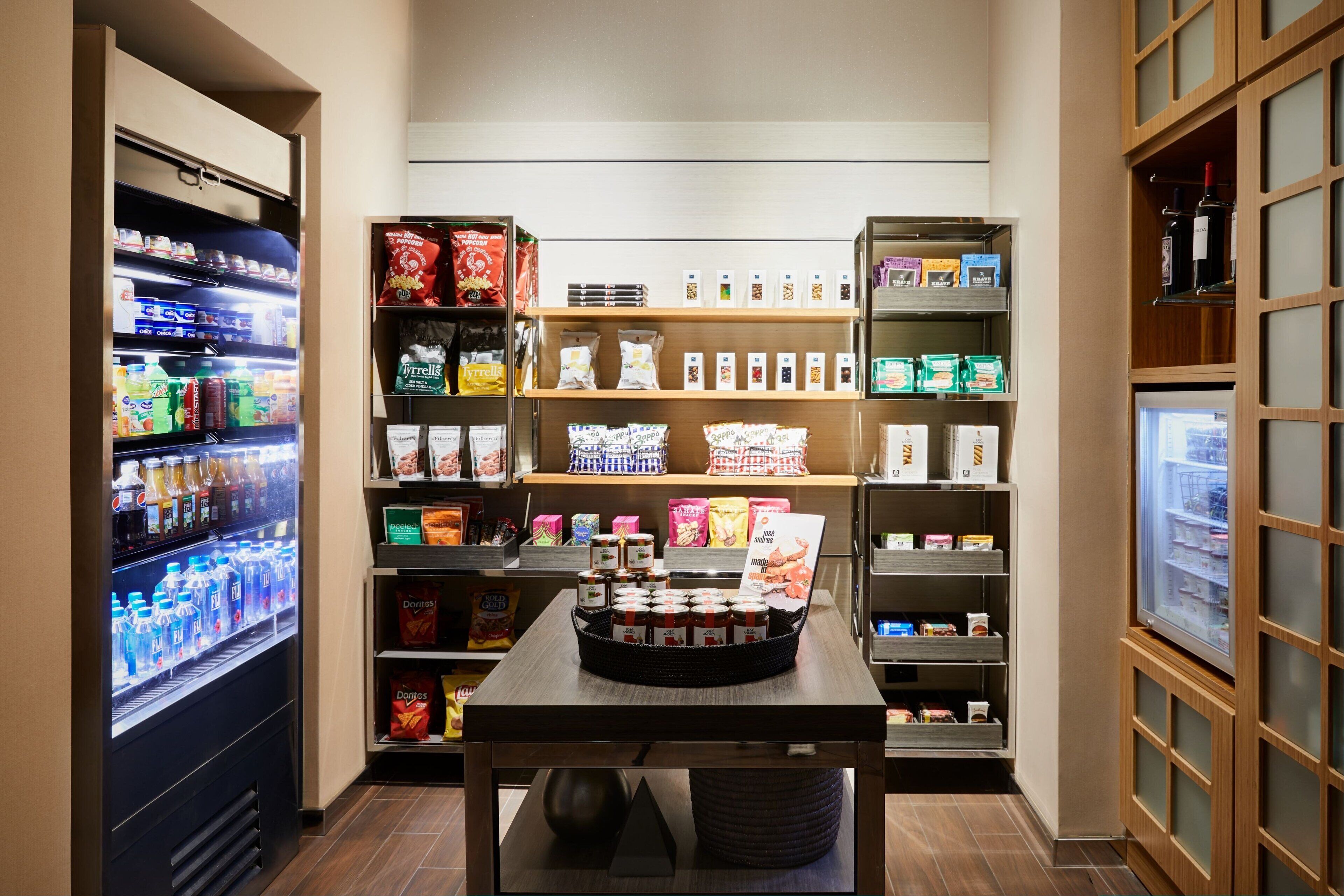 Interior de una tienda de comestibles con una sección refrigerada, estantes llenos de refrigerios y una mesa de exhibición con productos envasados.