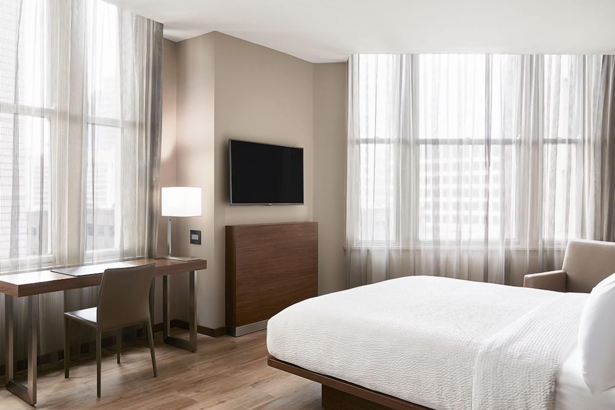 Une chambre d’hôtel avec un lit, un bureau et une chaise, une télévision et des rideaux en voile.