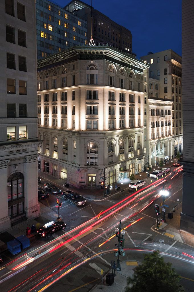 A historic building with classical architecture, a prominent corner entrance, and a street with light trails indicating traffic.
