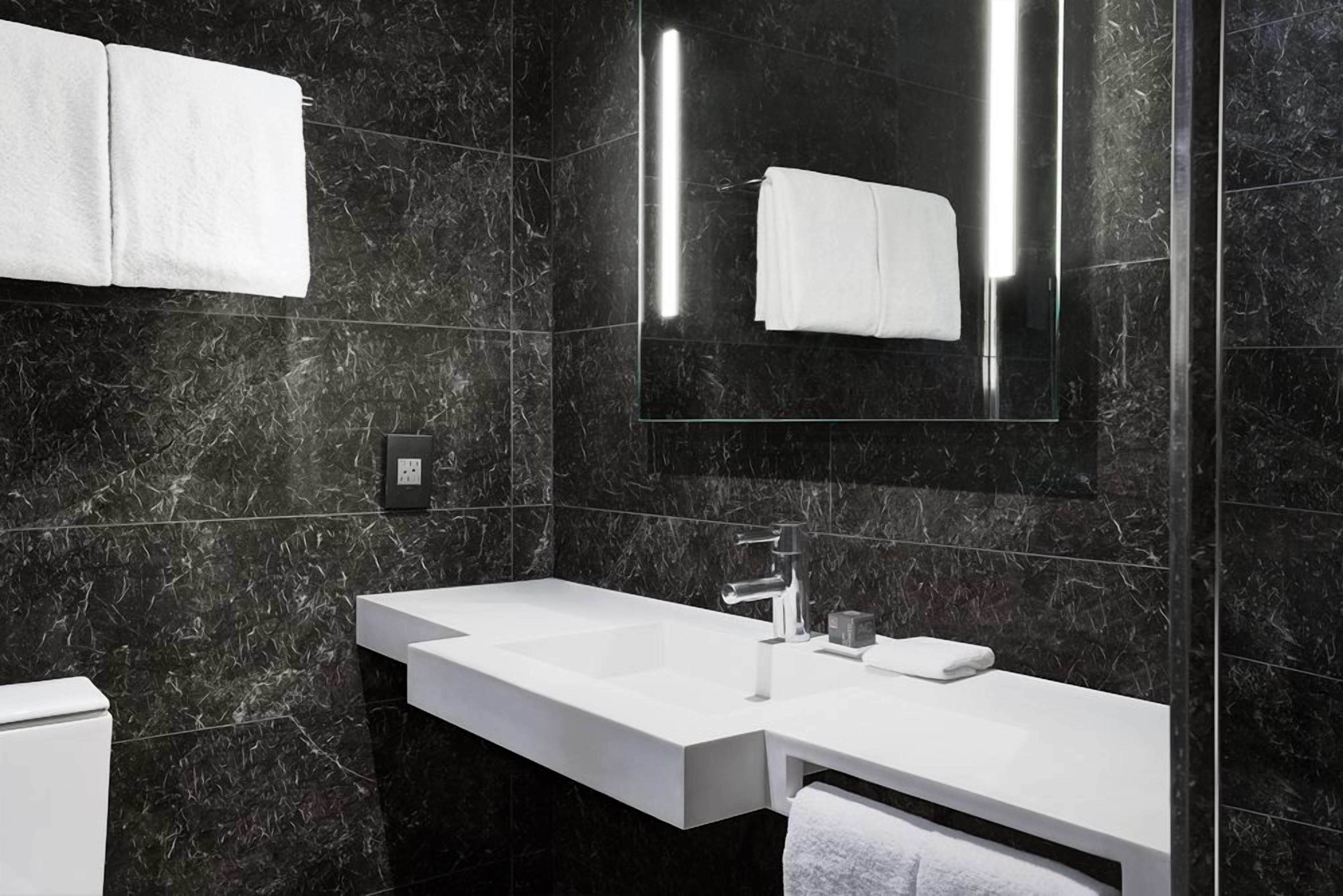 Un baño moderno con un lavamanos blanco, un espejo y paredes de mármol negro.