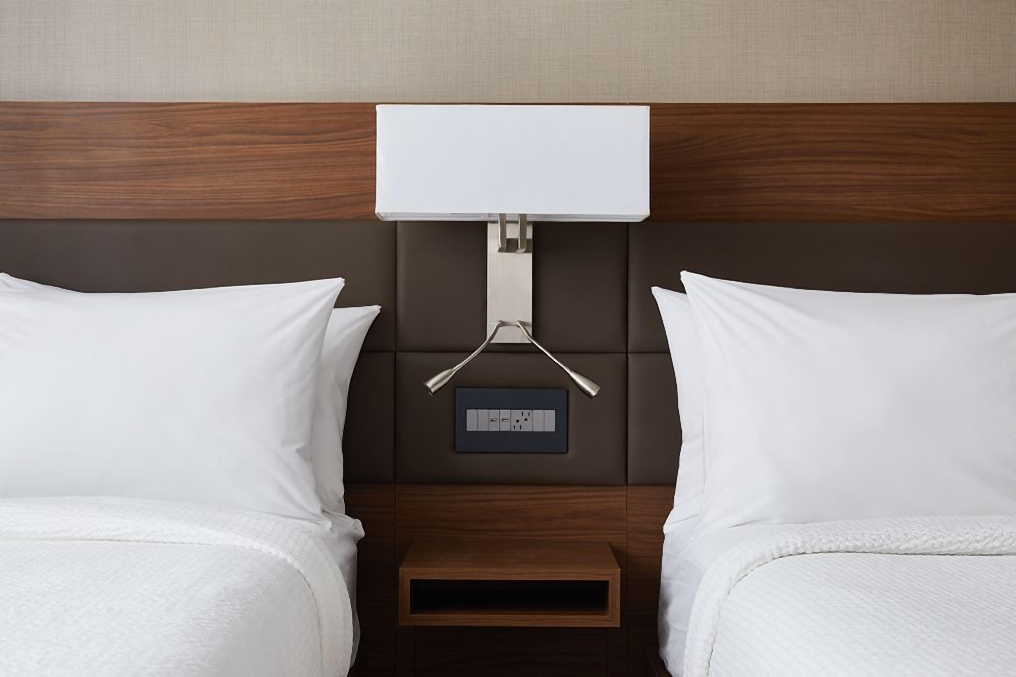 A hotel room with two beds, a bedside table, a wall-mounted power outlet, and a small shelf.
