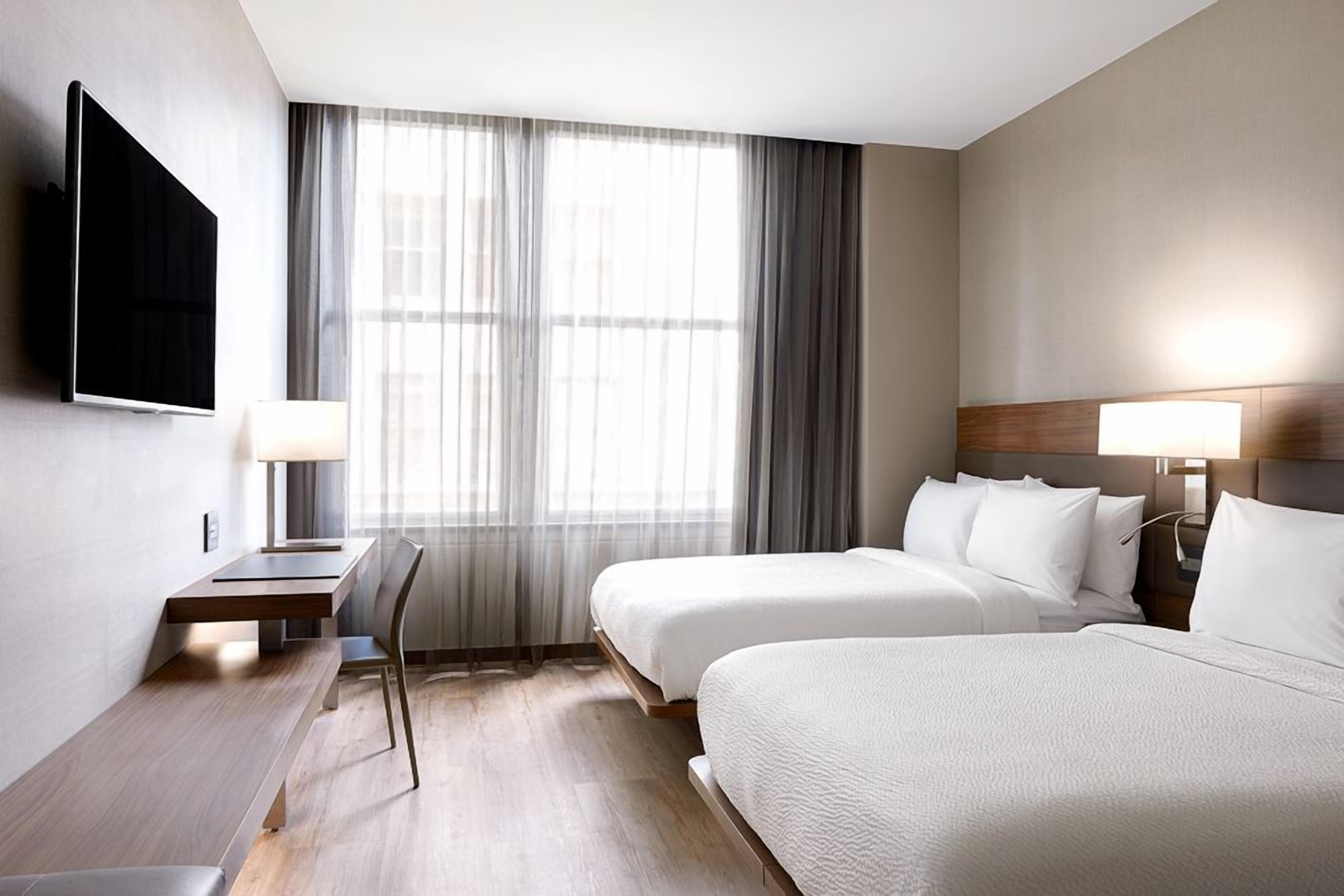 A hotel room with two beds, a desk, a TV, and a large window with curtains.
