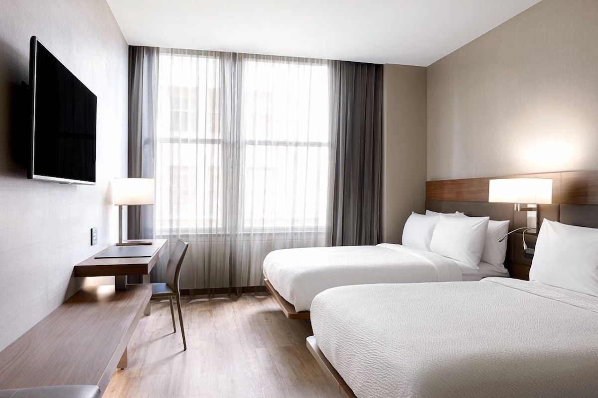 Une chambre d’hôtel avec deux lits, un bureau, une télévision et une grande fenêtre avec des rideaux.
