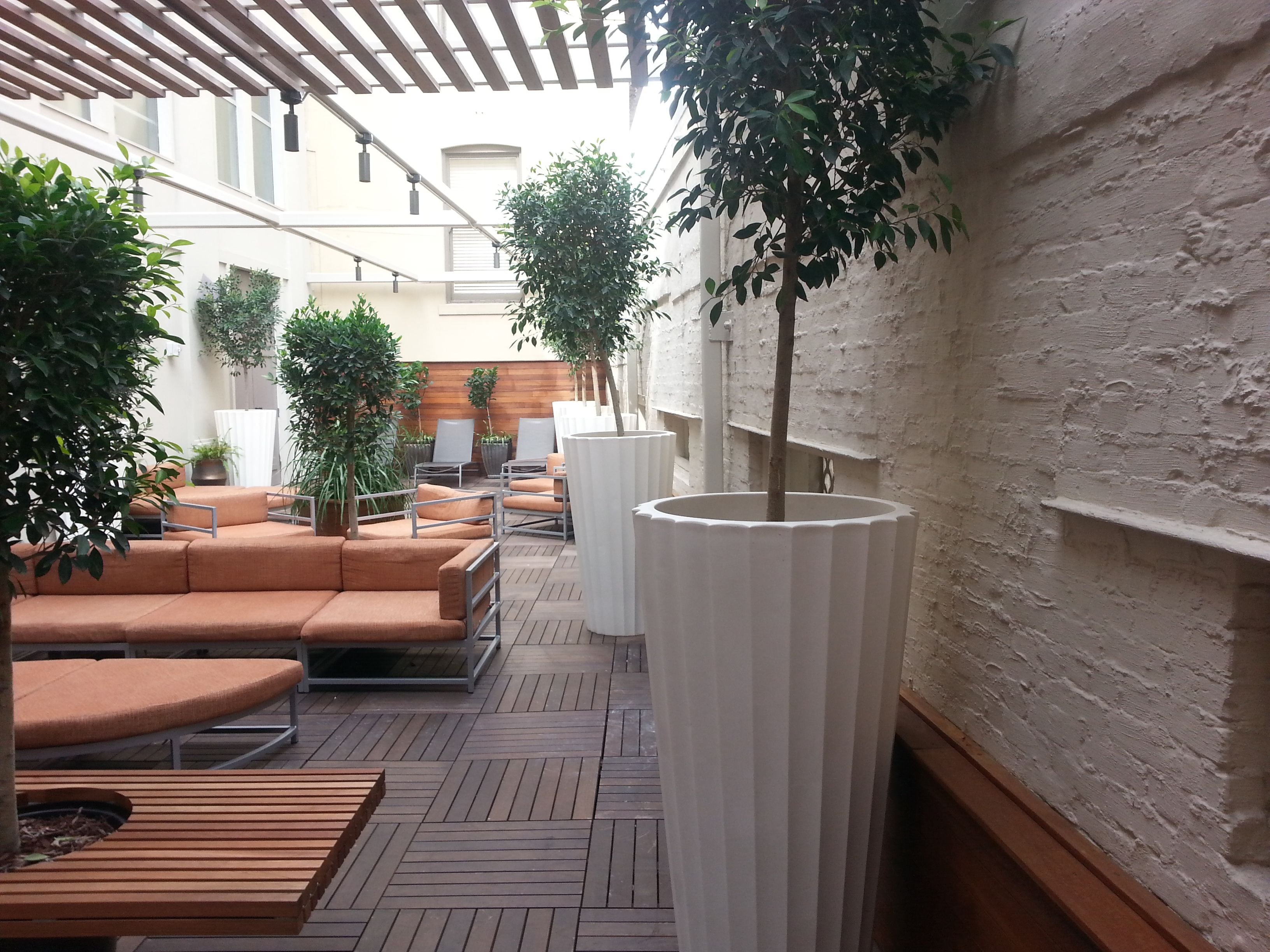 Een terras met houten banken en oranje kussens, omringd door potplanten en een witte kolomconstructie.