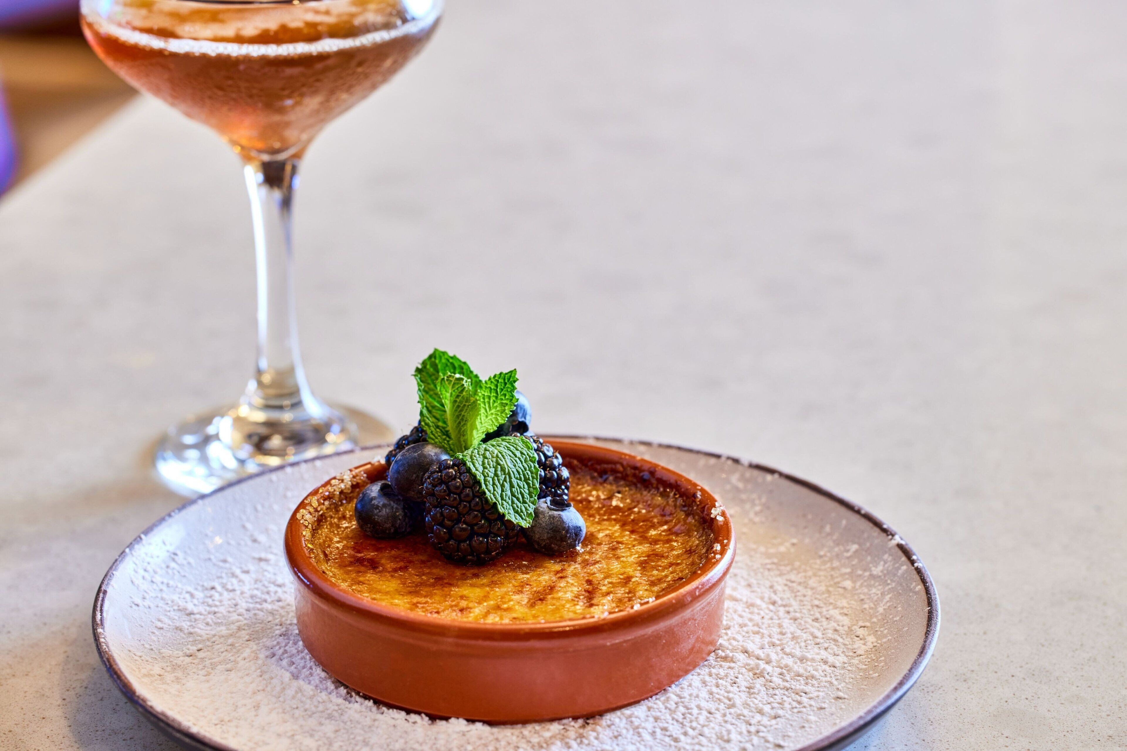 Un dessert servi dans un plat en céramique, posé sur une assiette, décoré de myrtilles et de menthe, avec un verre d’une boisson sombre en arrière-plan.