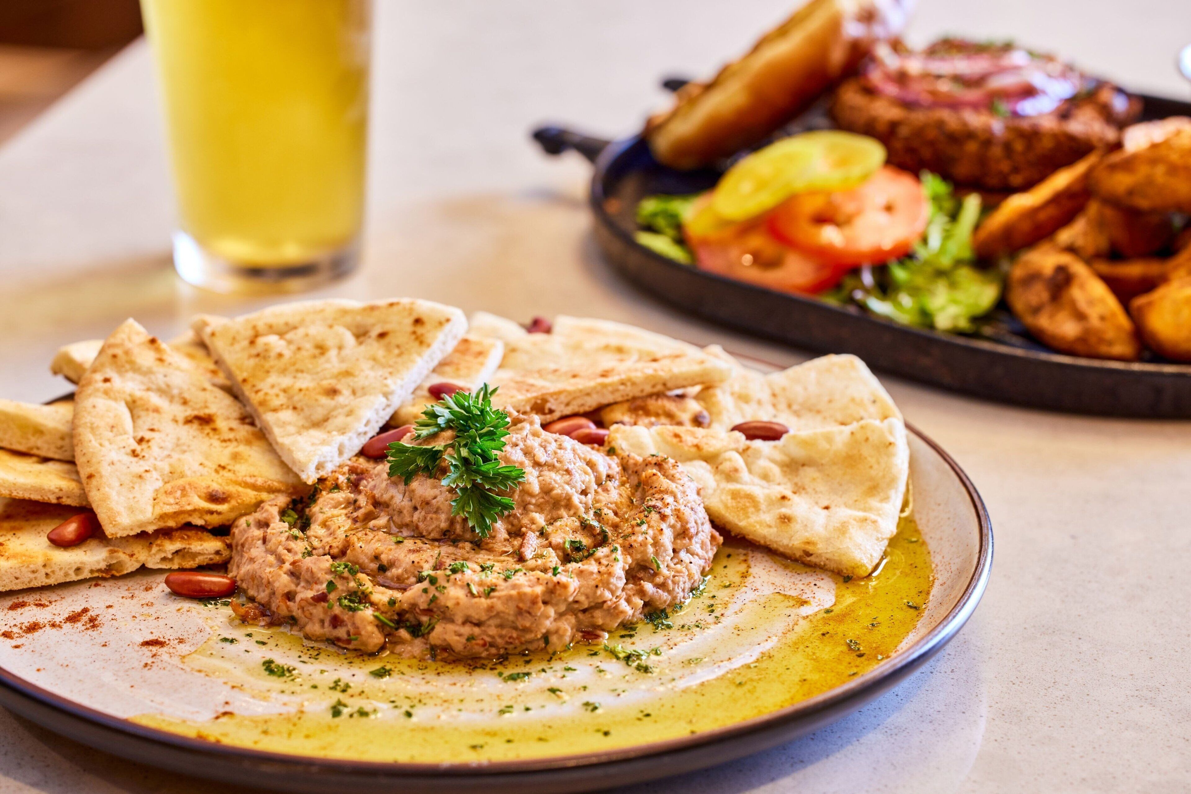 Un plato de hummus con pan de pita, una ensalada de tomates y pepinos, y un vaso de bebida amarilla sobre una mesa.
