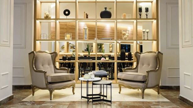 Un interior lujoso con dos sillones ornamentados, una mesa redonda central y una vitrina iluminada con diversos objetos decorativos.