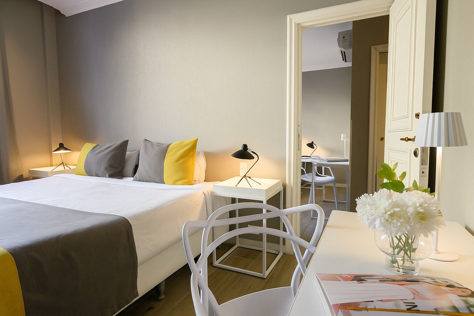A hotel room with a bed, bedside tables, a desk, a chair, and a vase with flowers.