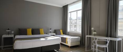 A modern hotel room with a large bed, two bedside tables, a desk with a chair, and a large window with curtains.