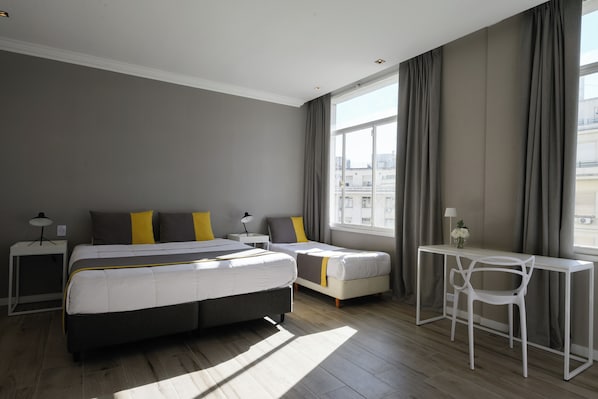 A modern hotel room with a large bed, two bedside tables, a desk with a chair, and a large window with curtains.