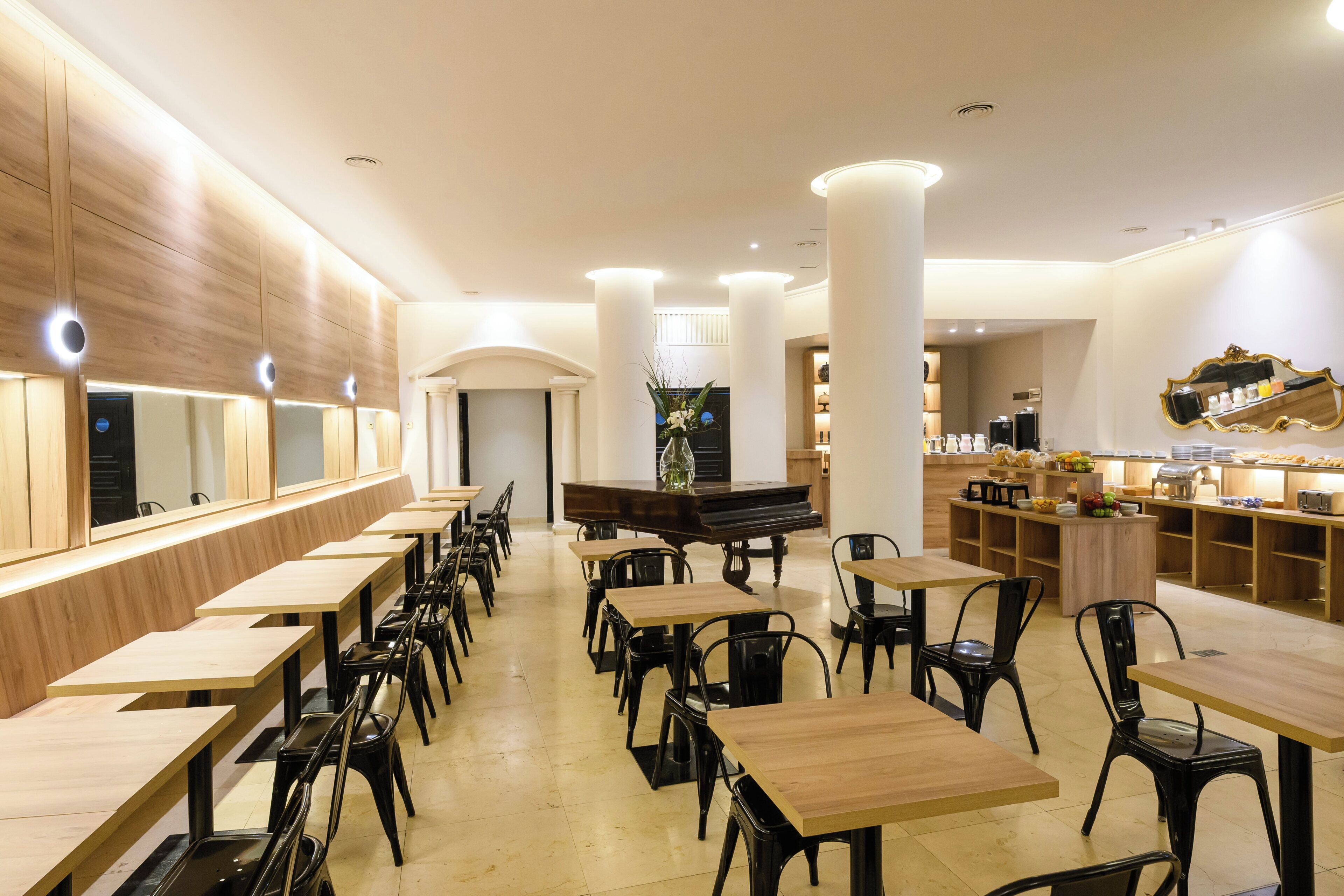 A restaurant with long wooden tables and black chairs, a piano, and a counter with various items.