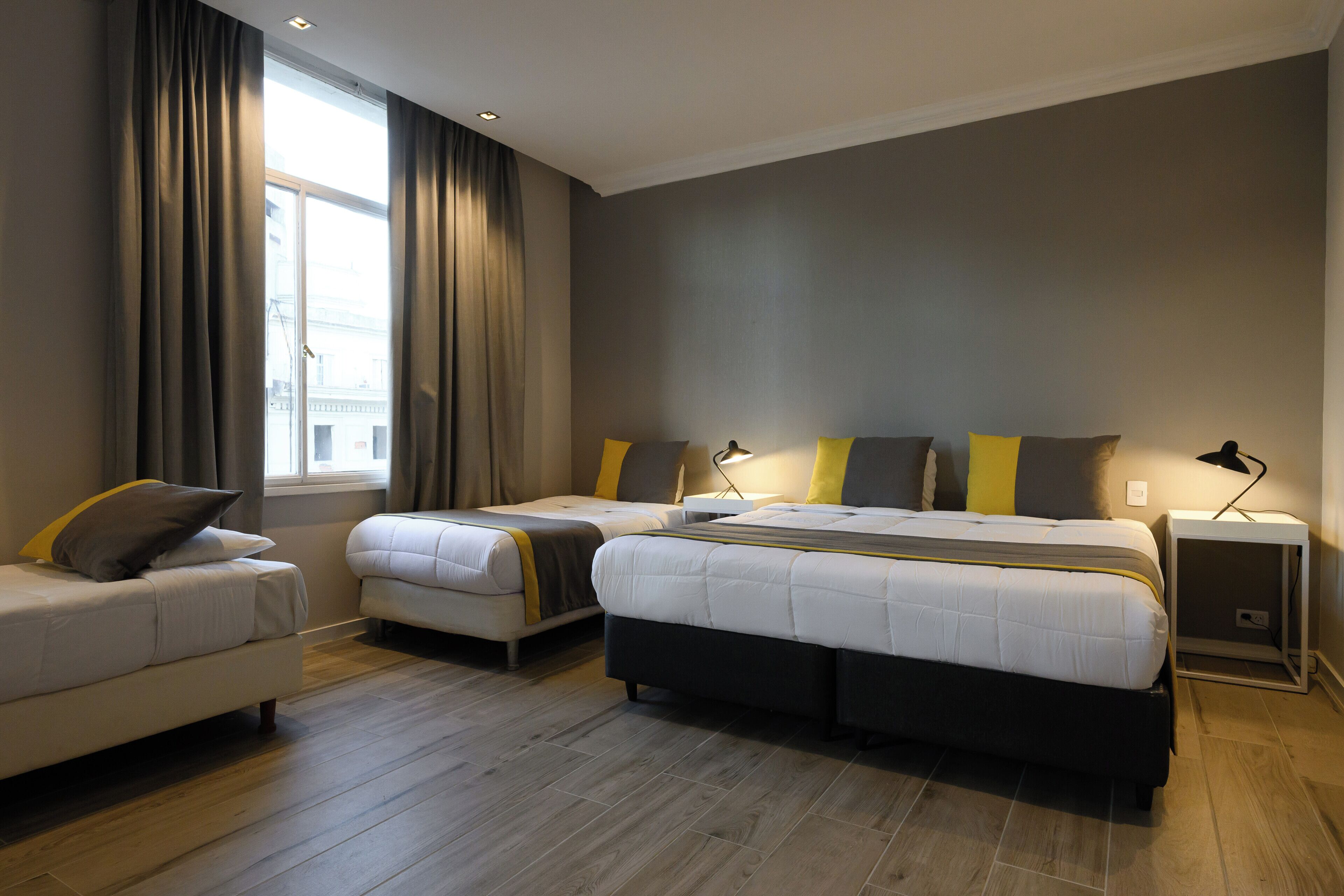 A modern hotel room with a large bed, two smaller beds, a sofa, and a window with curtains.