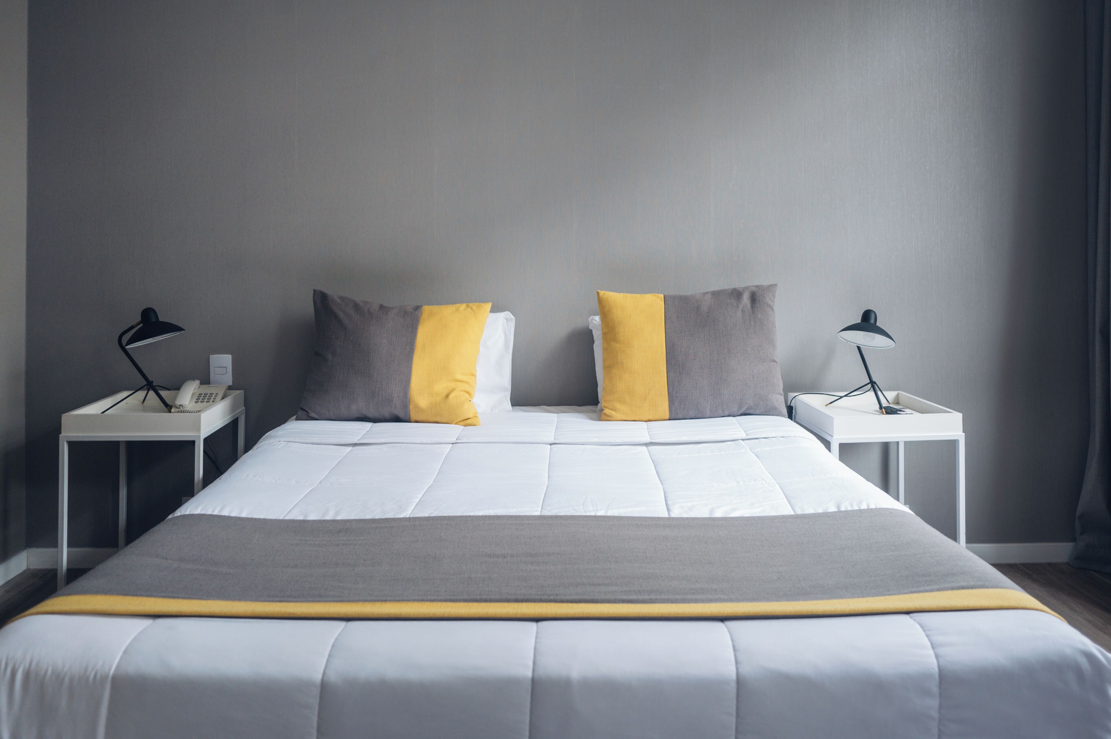 A neatly made bed with a white and gray duvet, two pillows with a yellow accent, and two bedside tables with lamps and a phone.