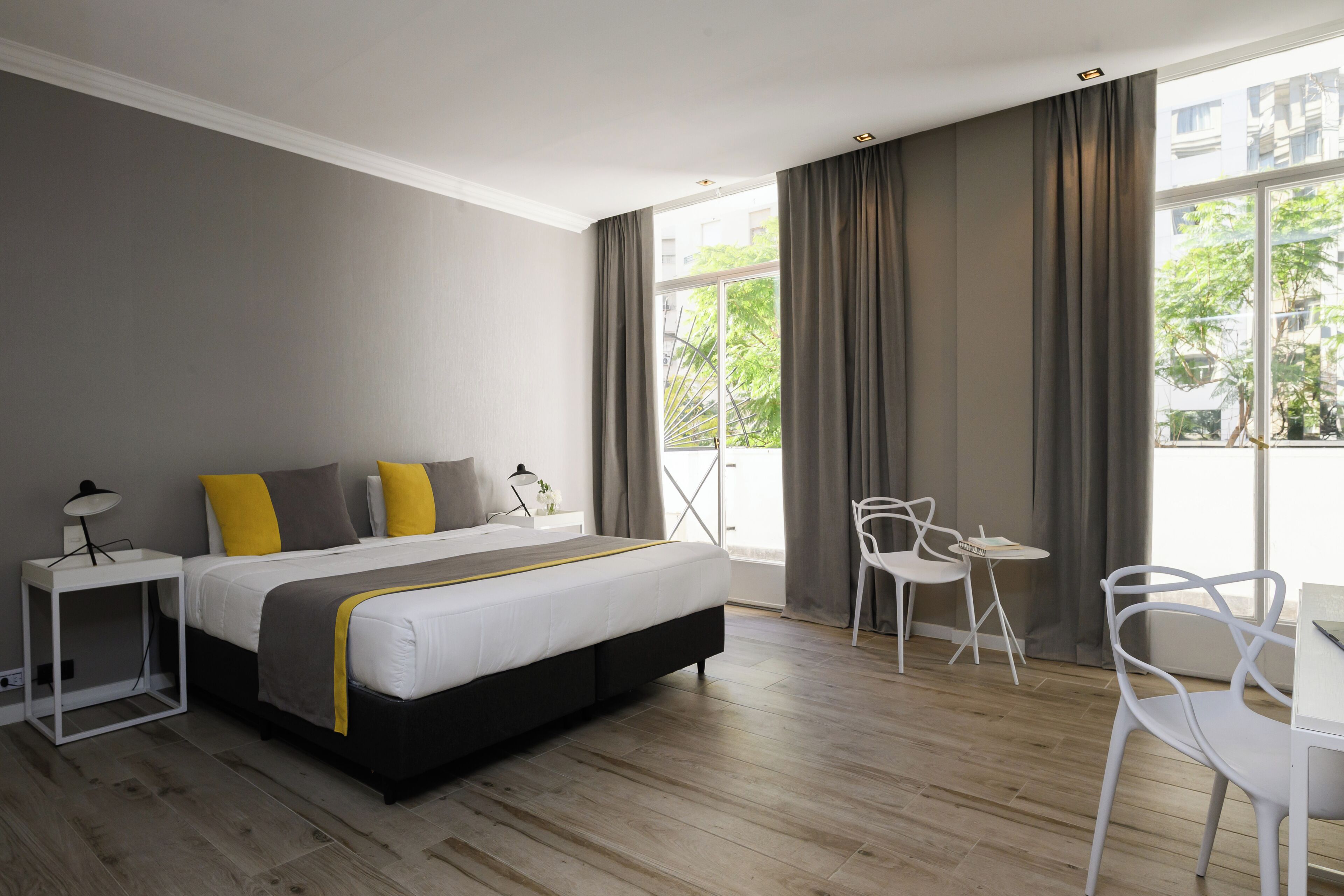 Habitación de hotel moderna con una cama grande, una mesita de noche con lámpara, una pequeña mesa con sillas y vistas al exterior a través de amplios ventanales.