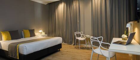 A modern hotel room with a large bed, a desk with a laptop, white chairs, and a curtain backdrop.