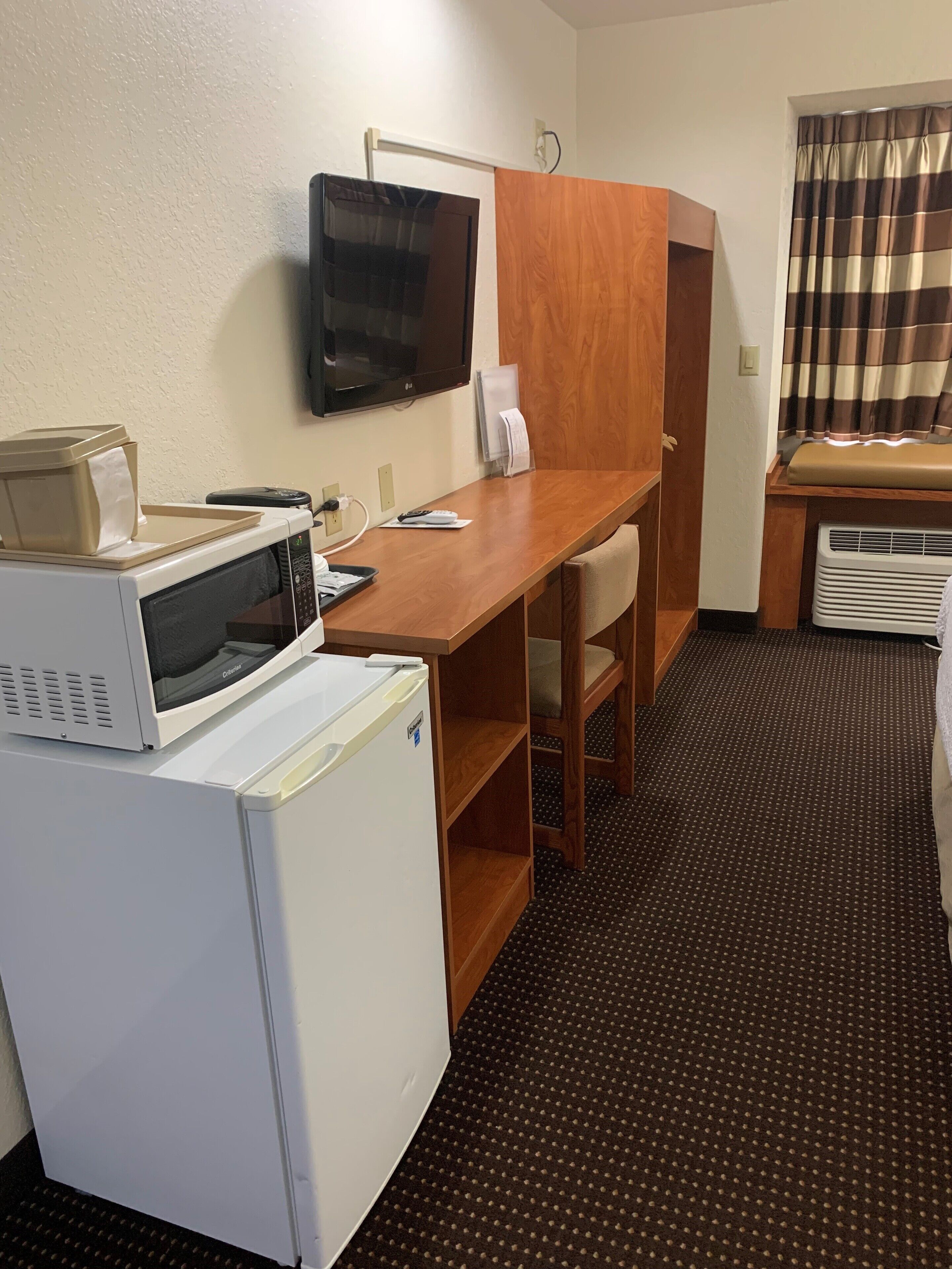Hotellihuone, jossa on pieni keittonurkkaus, johon kuuluu mikroaaltouuni, pieni jääkaappi, puinen työpöytä ja seinälle asennettu televisio.