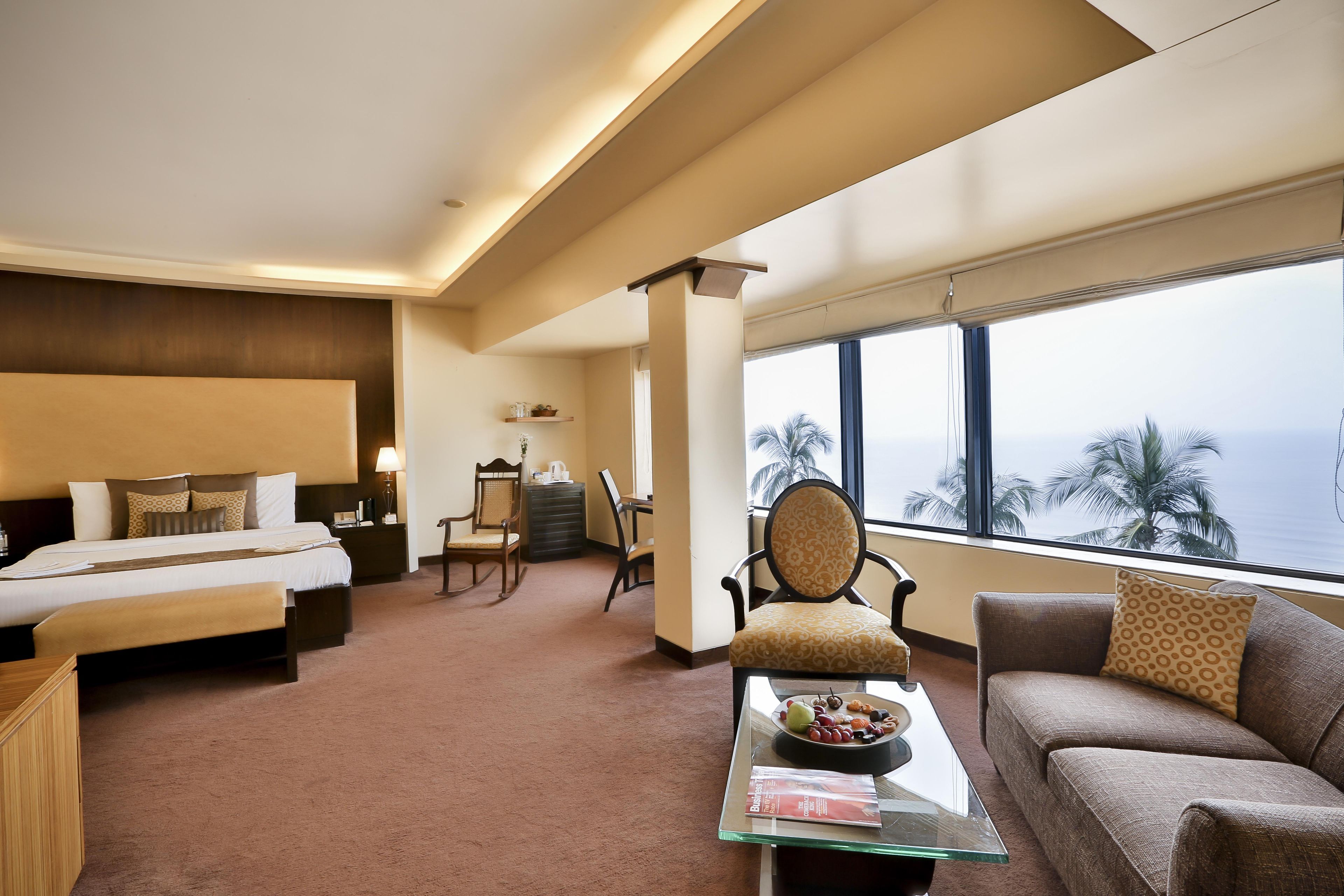 Ein Hotelzimmer mit einem großen Bett, einer Couch, einem Esstisch mit Obst und Blick aufs Meer.
