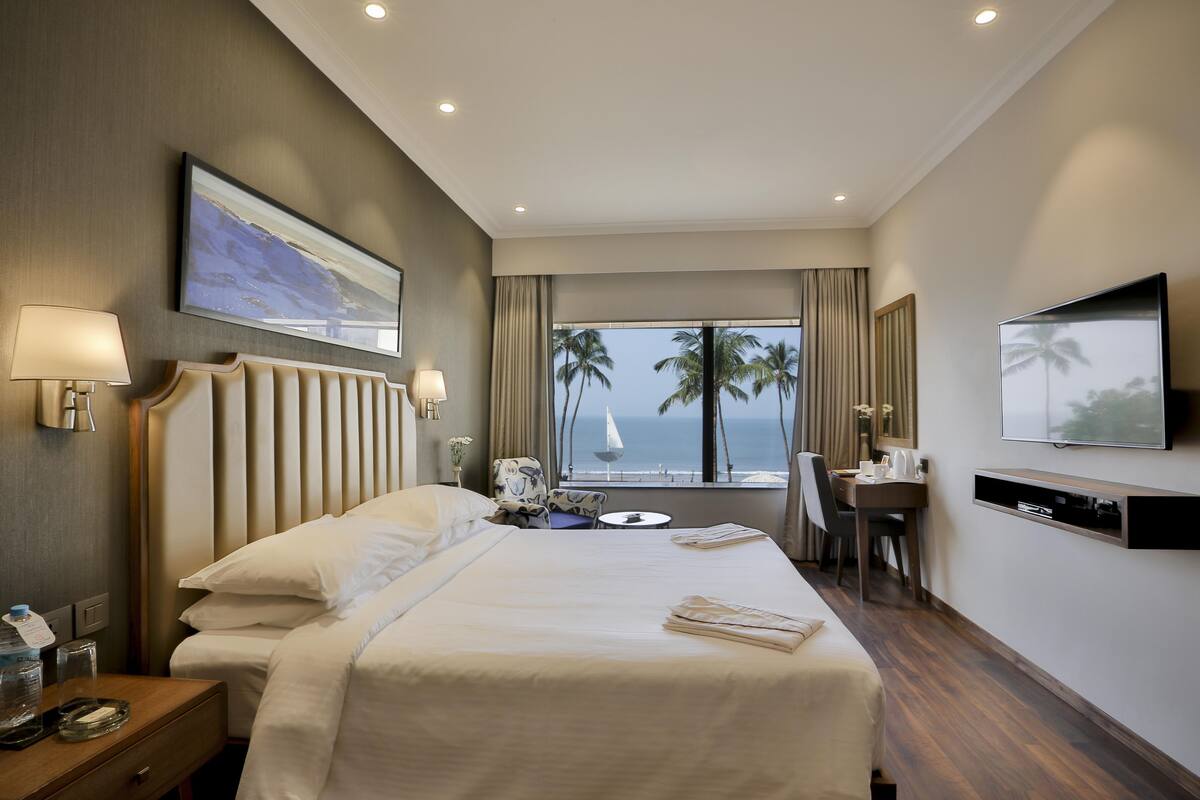 Ein Hotelzimmer mit einem großen Bett, einem Schreibtisch, einem Stuhl, einem Fernseher und Blick auf Palmen sowie das Meer.