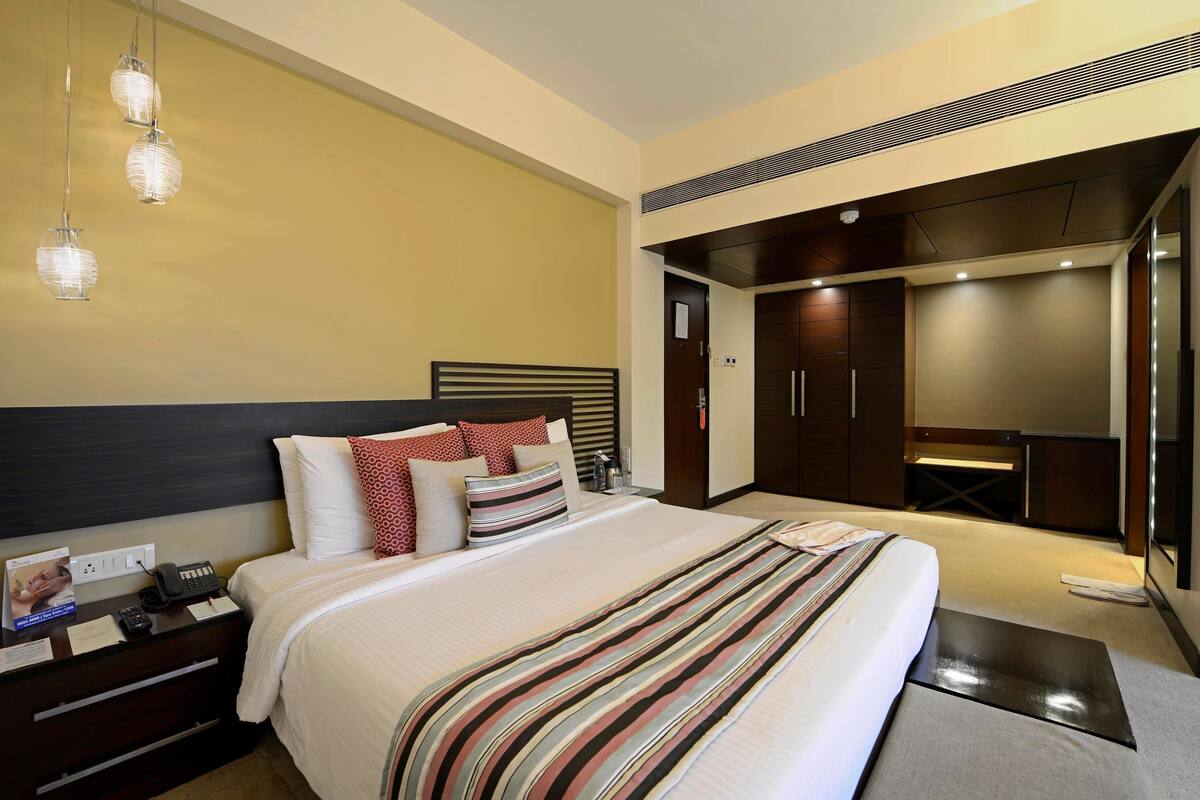 Ein modernes Hotelzimmer mit einem großen Bett, einem Nachttisch mit Telefon und Broschüre, einem Kleiderschrank und einem Schreibtisch mit Stuhl.