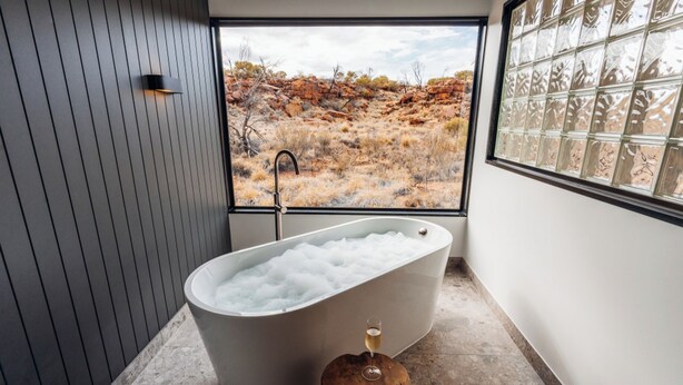 A modern bathroom with a freestanding bathtub, a wooden stool, and a large window with a desert view. - Discovery Resorts - Kings Canyon (Petermann)