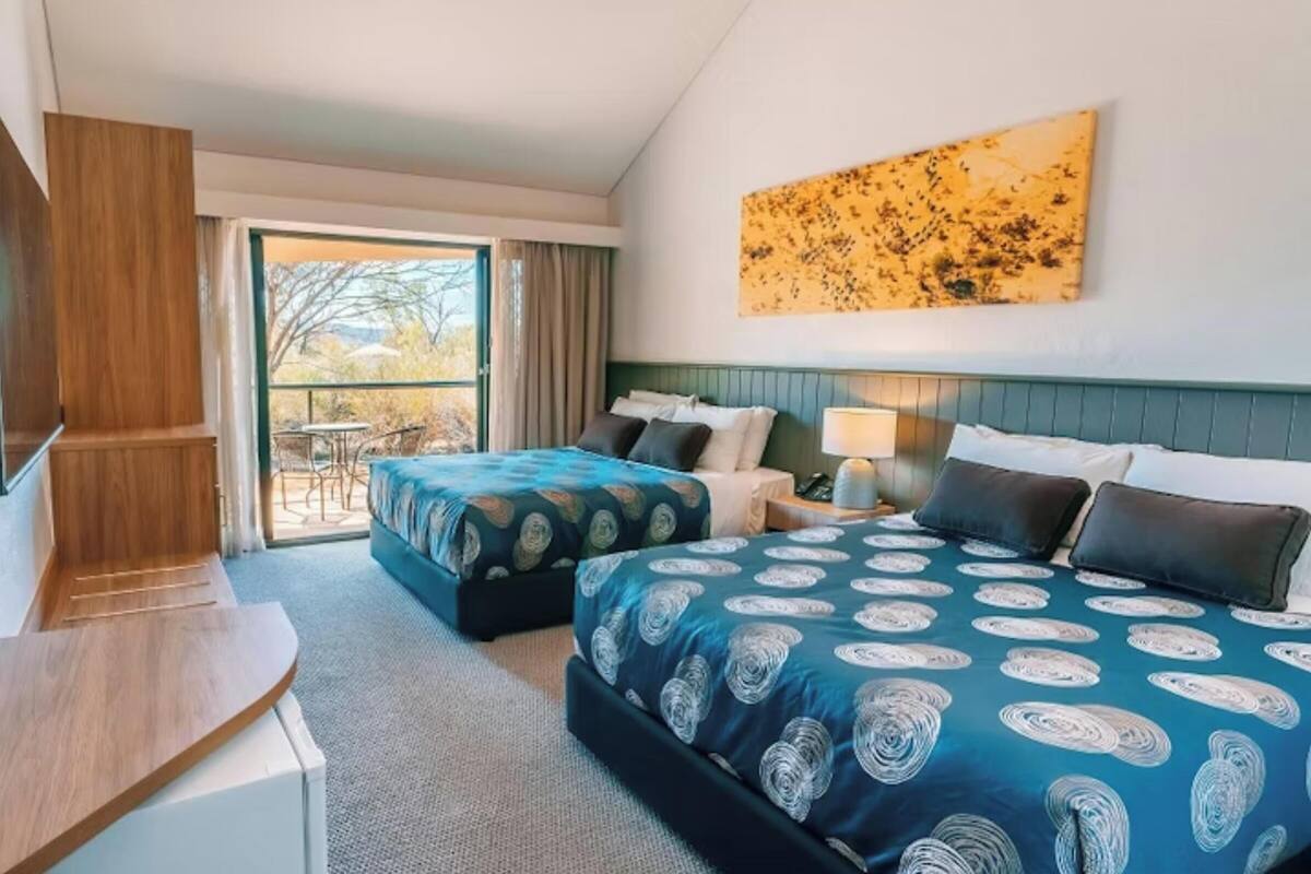 A hotel room with two beds, a desk, a TV, and a balcony with a table and chairs.