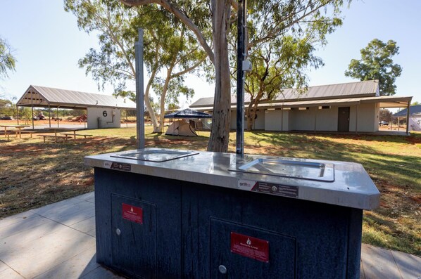 A portable grill station with a metal roof and a built-in cooler, surrounded by trees and a picnic table. - Discovery Resorts - Kings Canyon (Petermann)